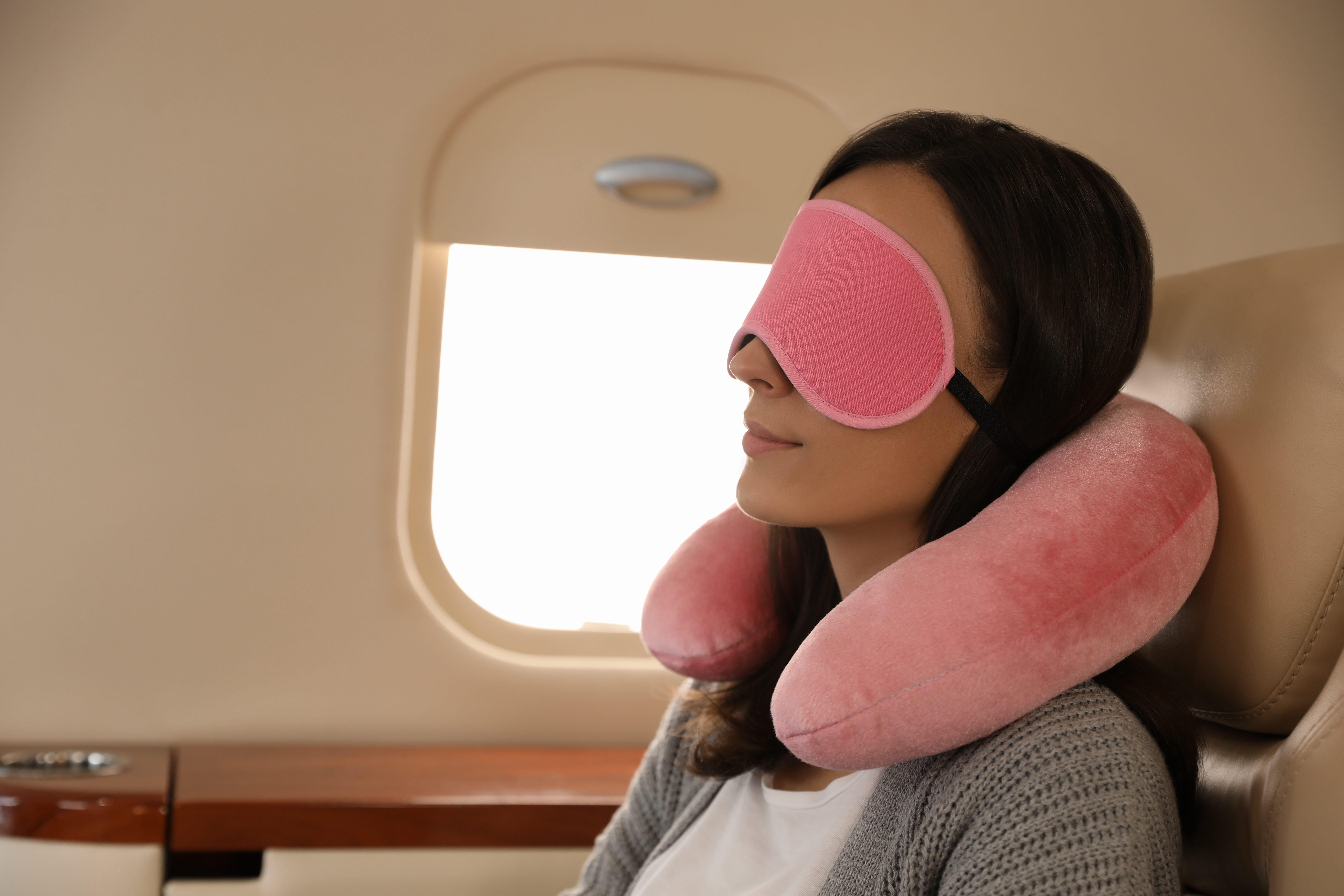 A woman asleep on a plain with a pink eye mask over her eyes and a matching pink neck pillow positioned between her shoulders and her chin