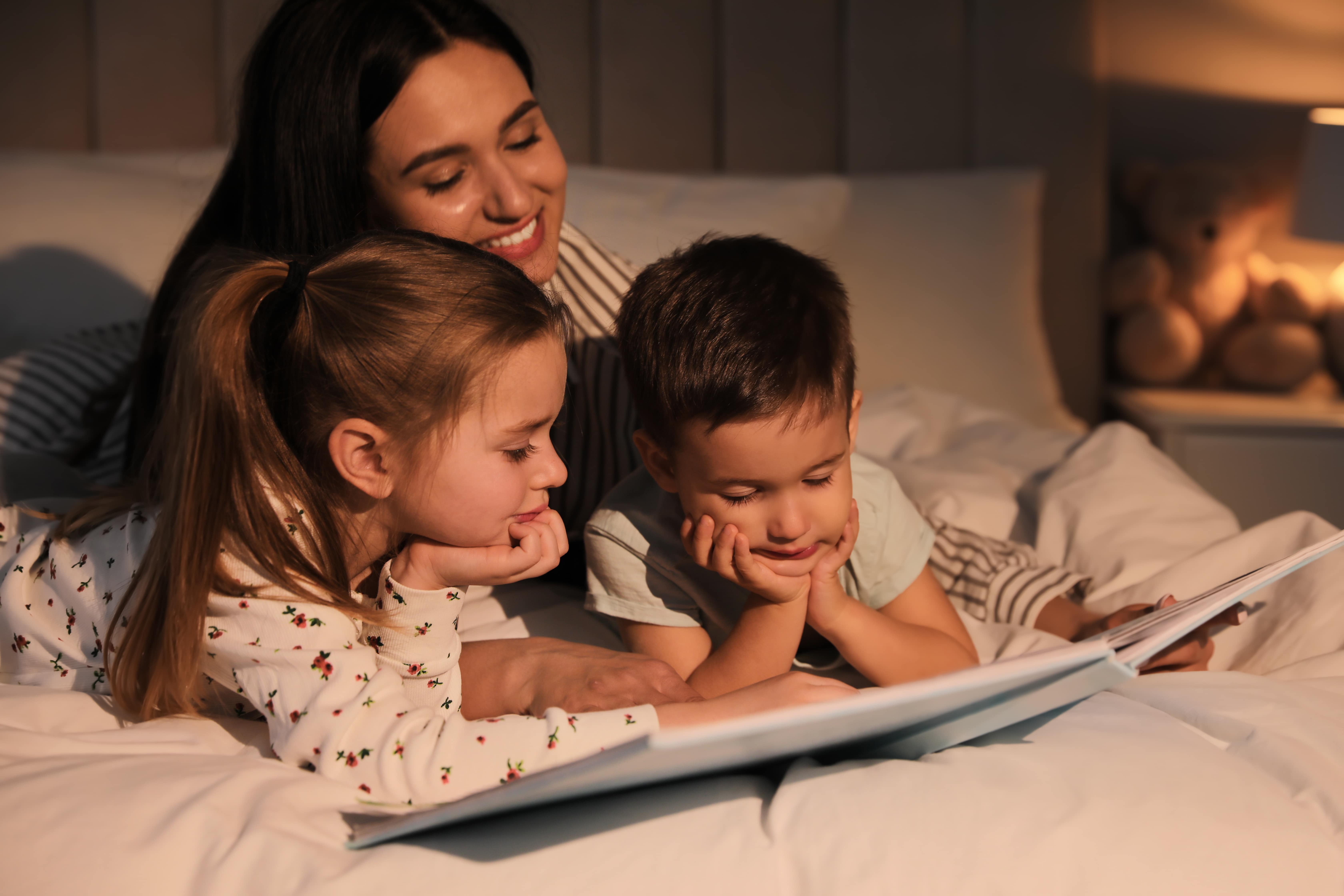 A mum reading to her two children in bed. Both are snugged in, looking at the pictures in the book as the mum reads and a teddy bedside a lamp is visible in the background