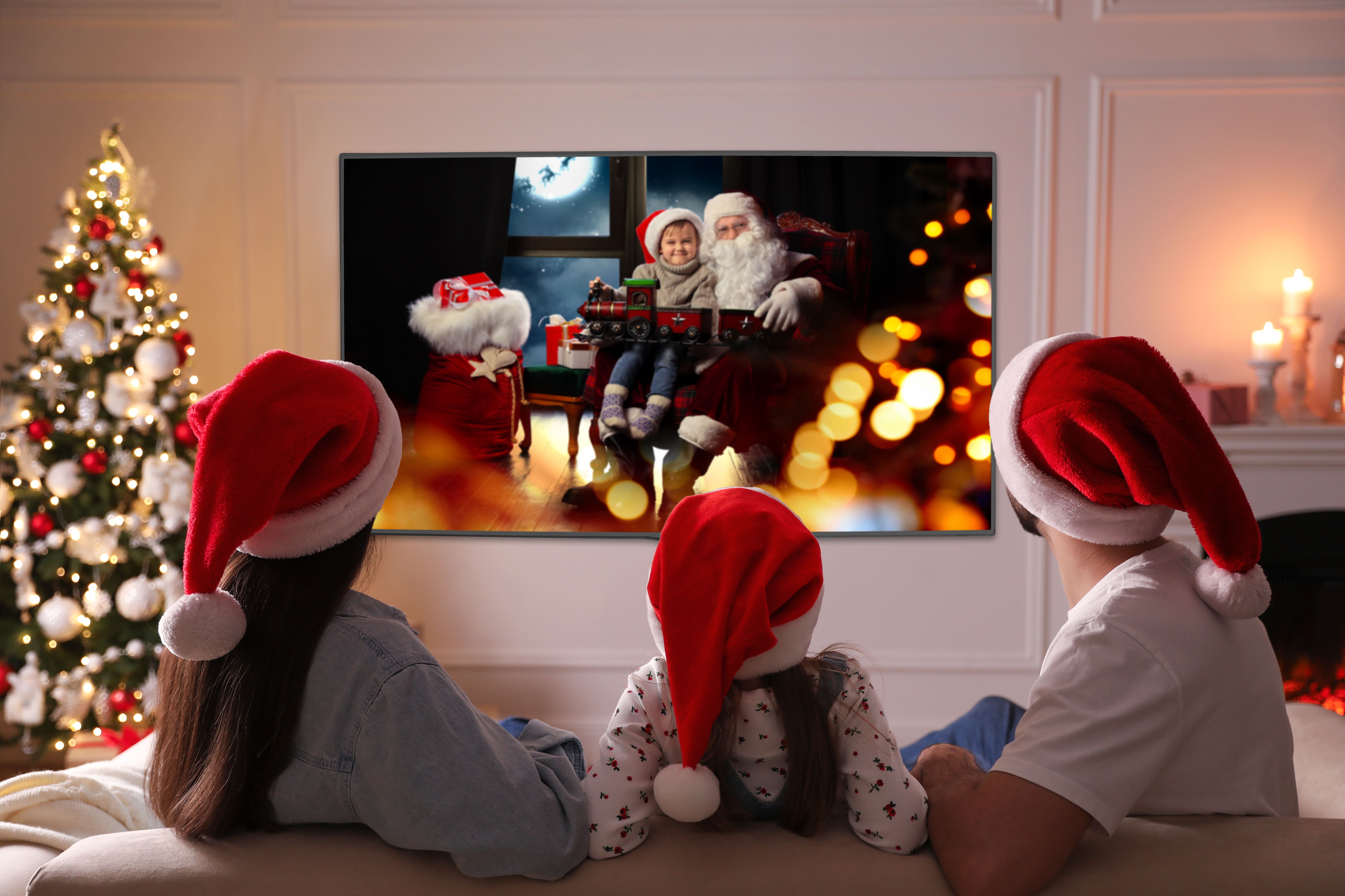 A family of three wearing Santa hats and snuggled close together while watching a Christmas film. The Christmas tree is twinkling with fairy lights beside the TV.
