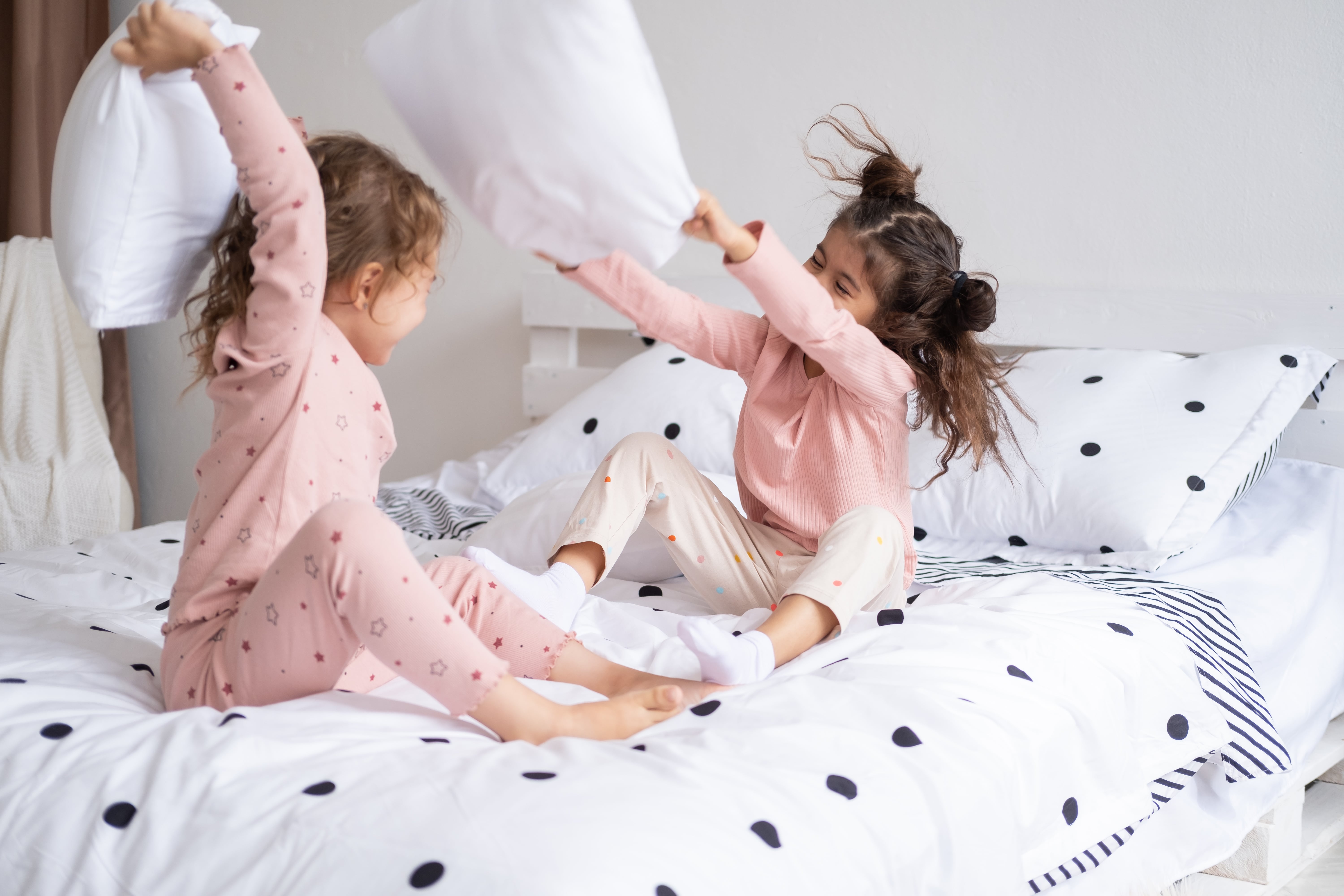 Two little girls in pink pyjamas having a pillow fight on a single bed dresses with a white and black polkadot bedding set.