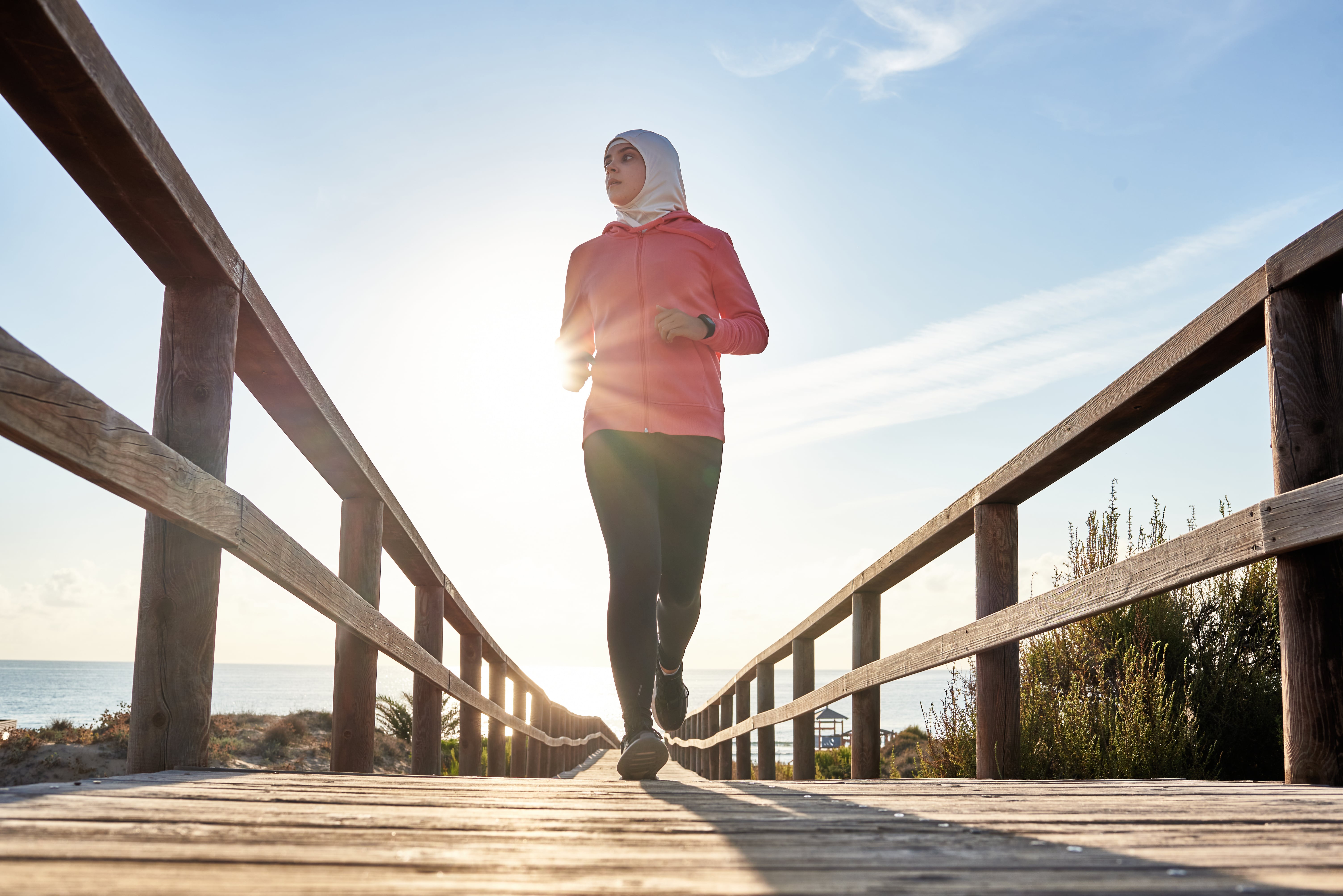 Woman wearing a head covering and athletic clothing jogs across a wooden bridge with the sea and countryside behind her.
