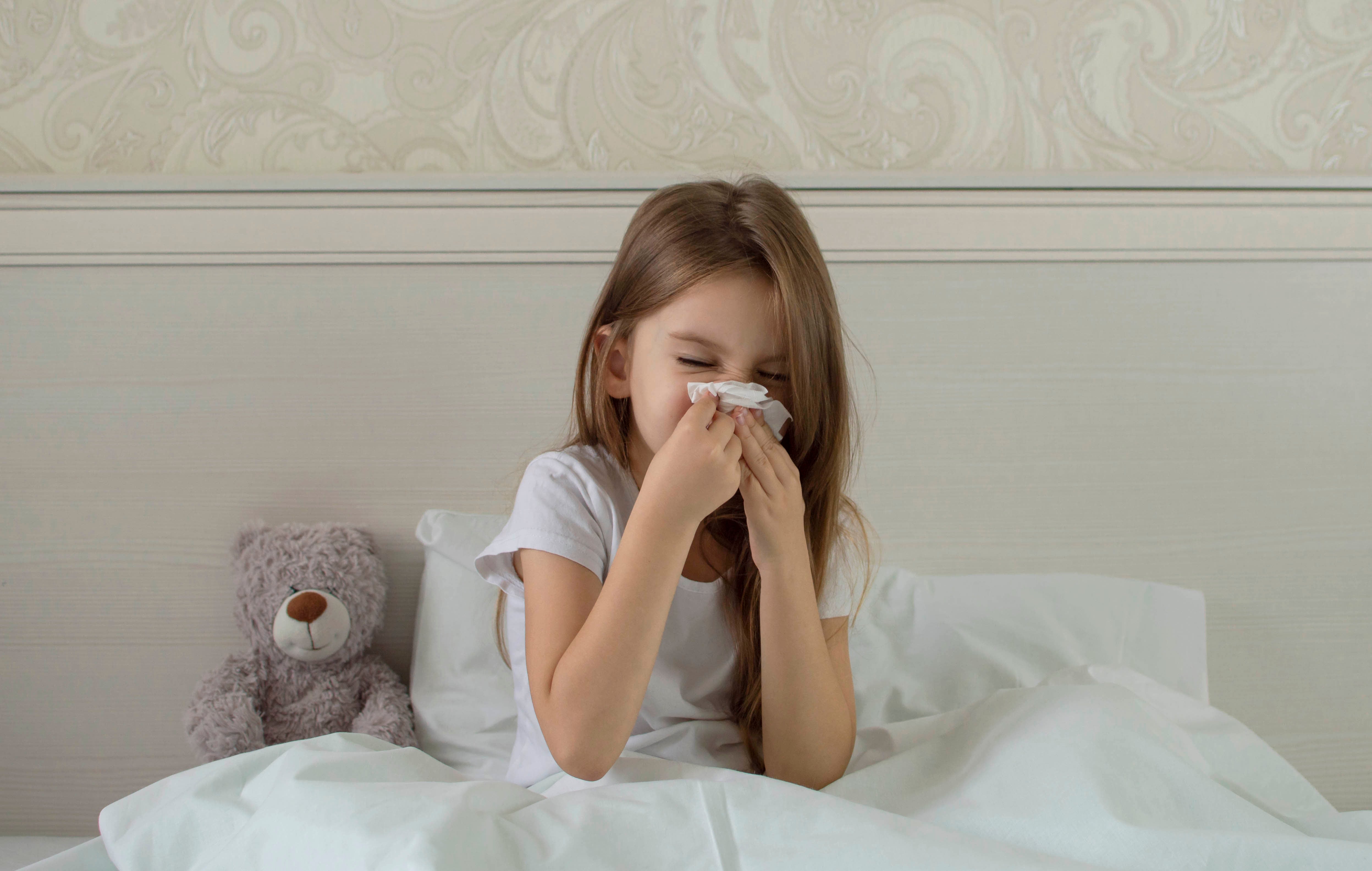 Young girl in bed blowing her nose with a tissue. A teddy bear sits next to her.