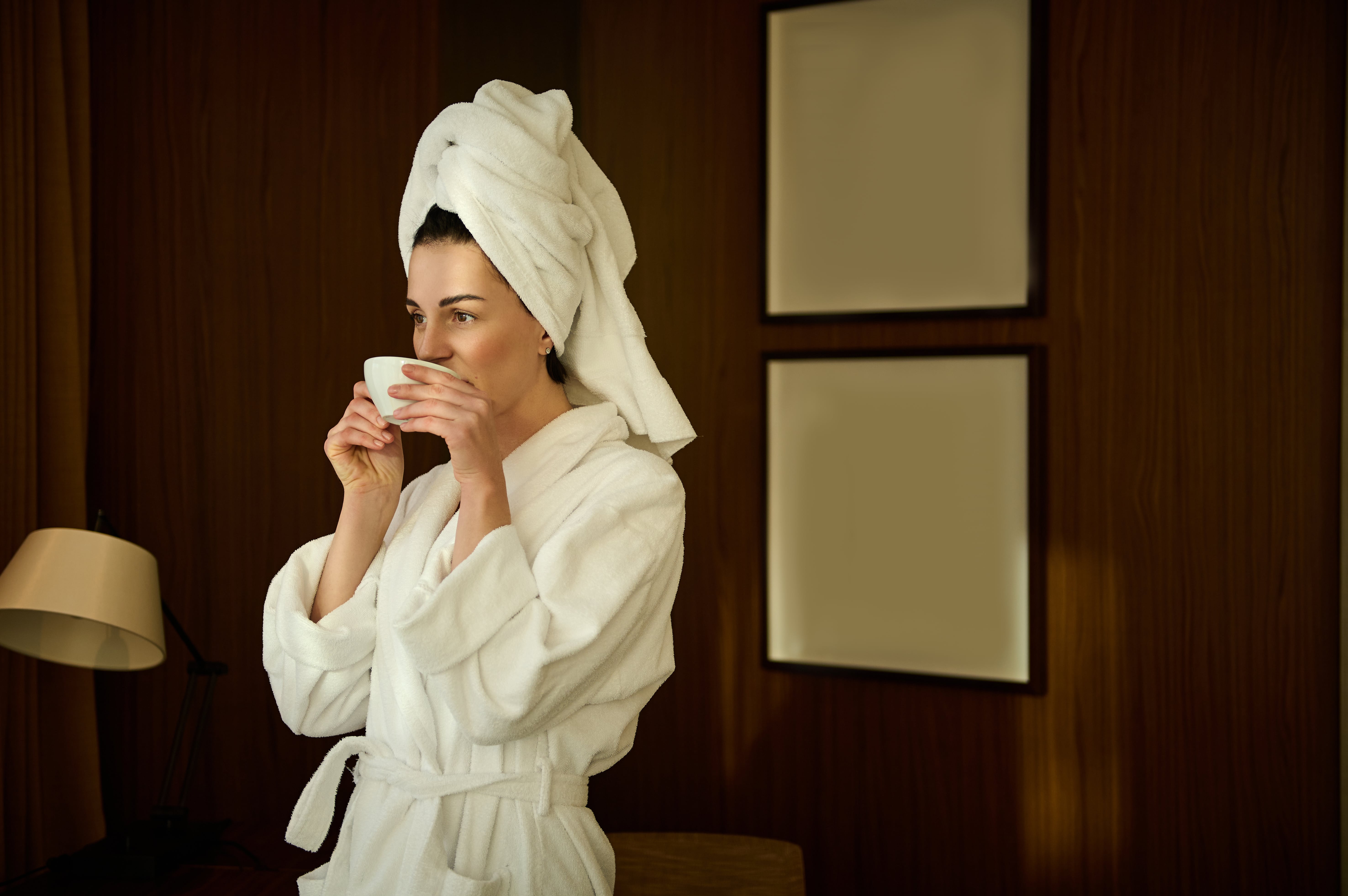 Woman in  bathrobe with head wrapped in a towel drinking from a cup.