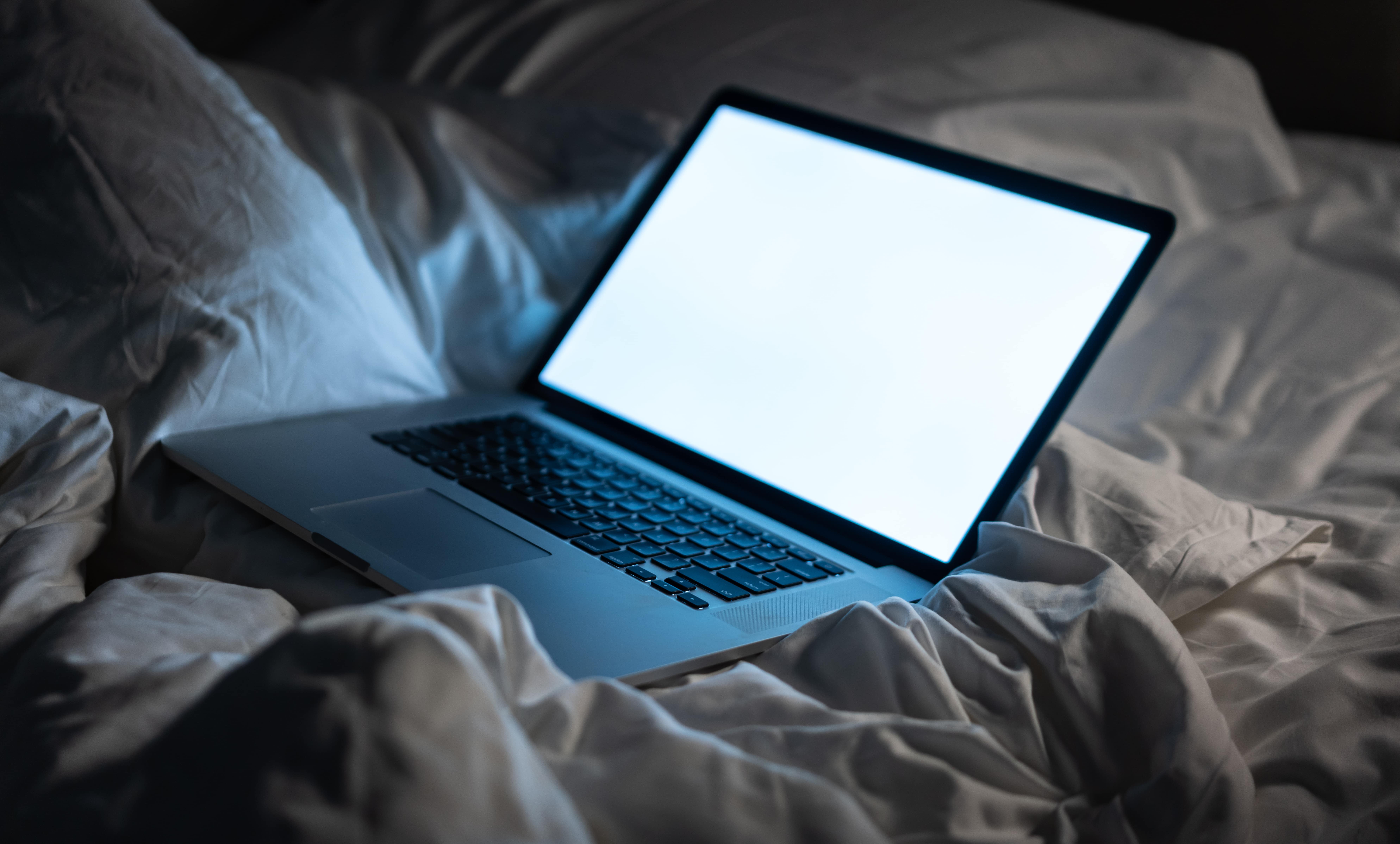 An open laptop with bright blank screen sits on a dishevelled bed.