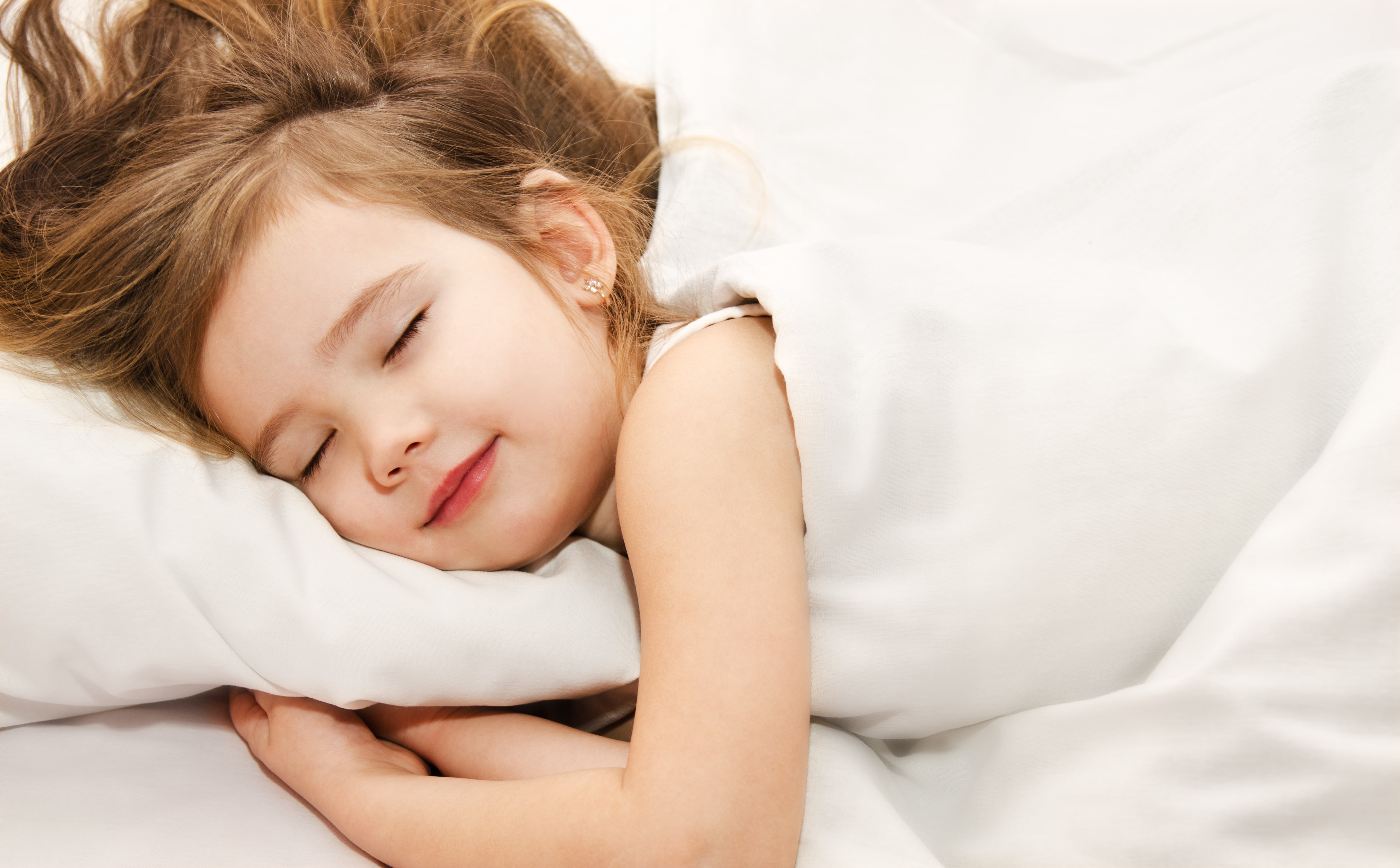 A little girl sleeping under a white duvet, hugging her pillow and smiling while her hair cascades around her 