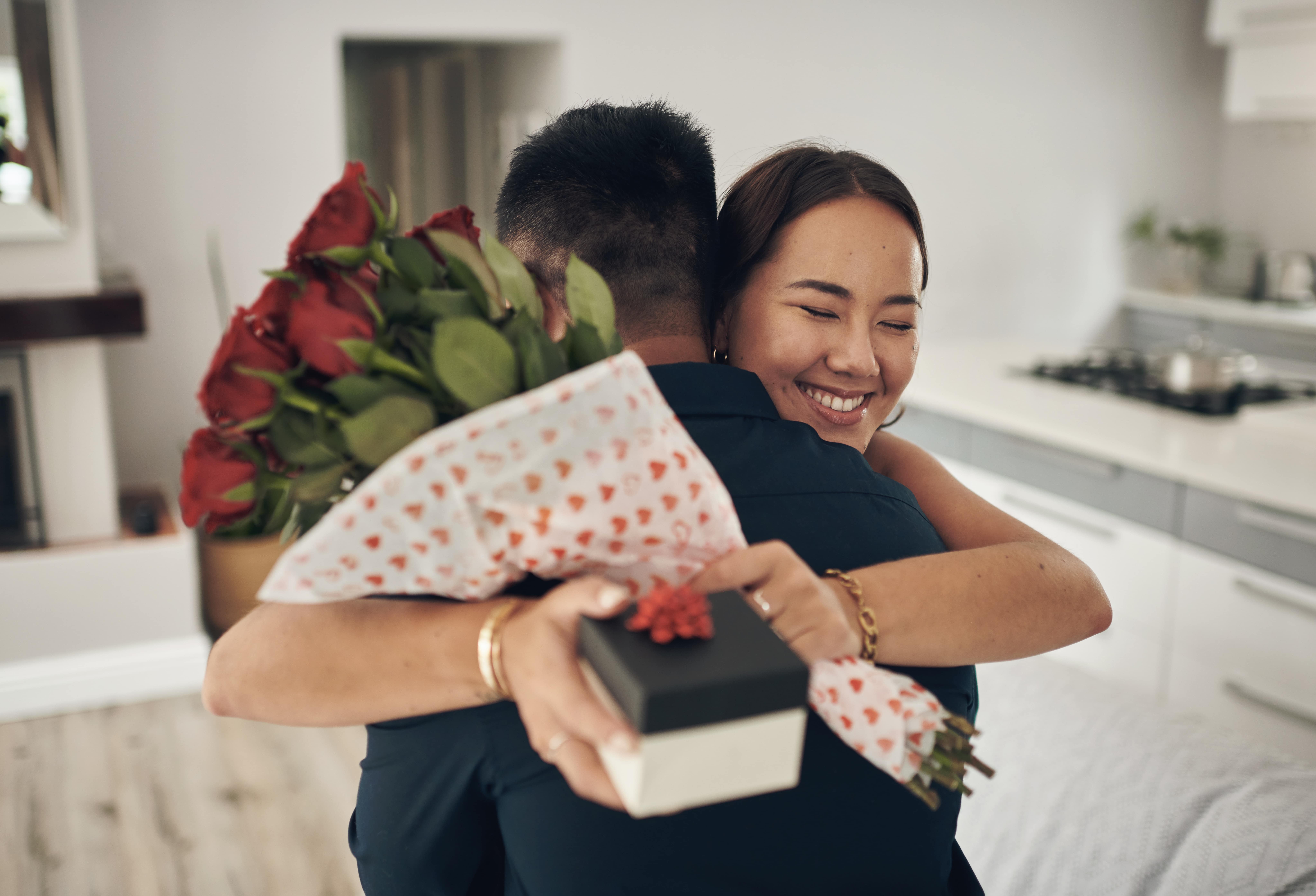 A woman hugs a man and she is holding a bouquet of red roses and a small box with a bow.