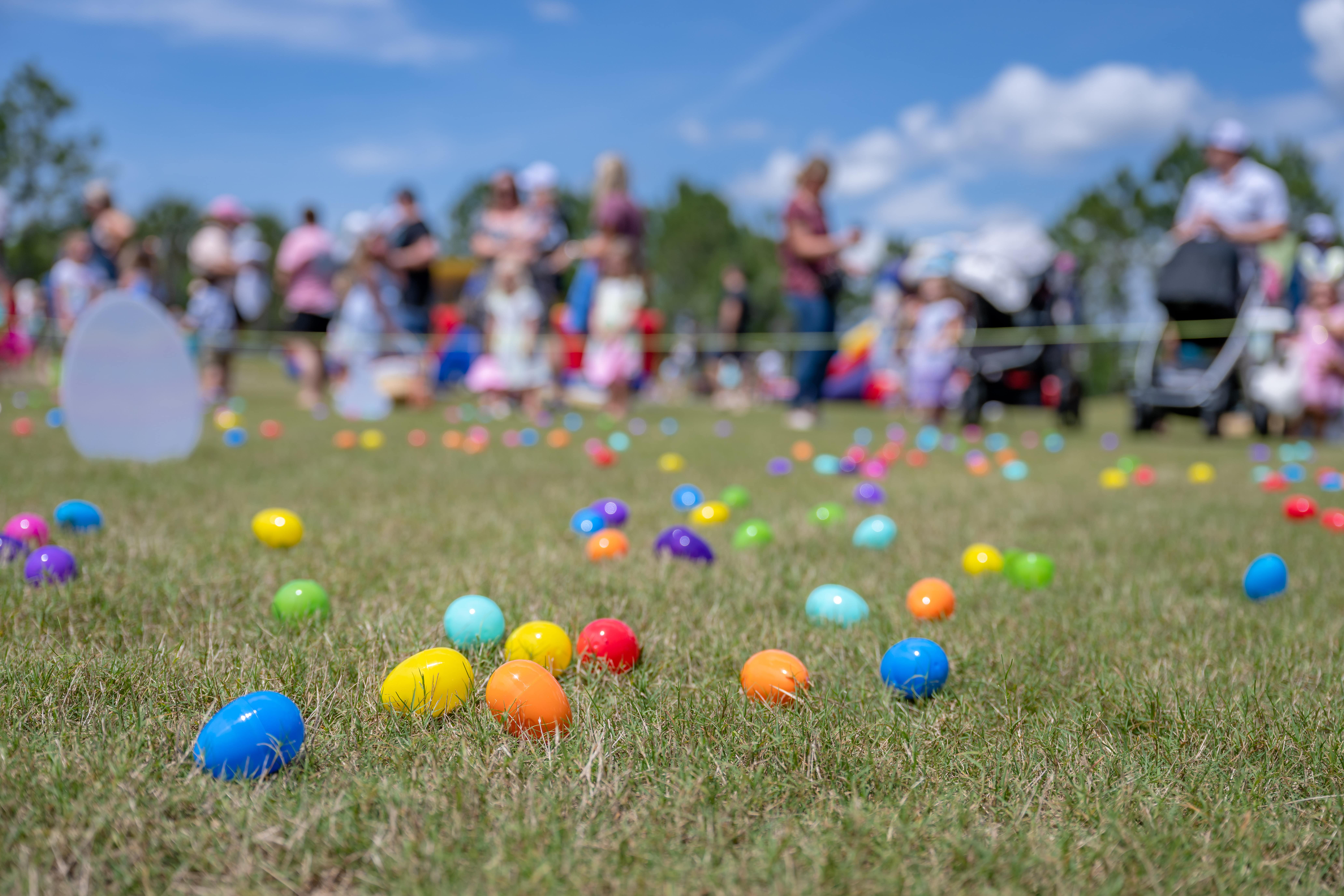 A field filled with brightly coloured Easter eggs scattered over the grass in the background is a line of children and parents with prams waiting to begin their Easter hunt.
