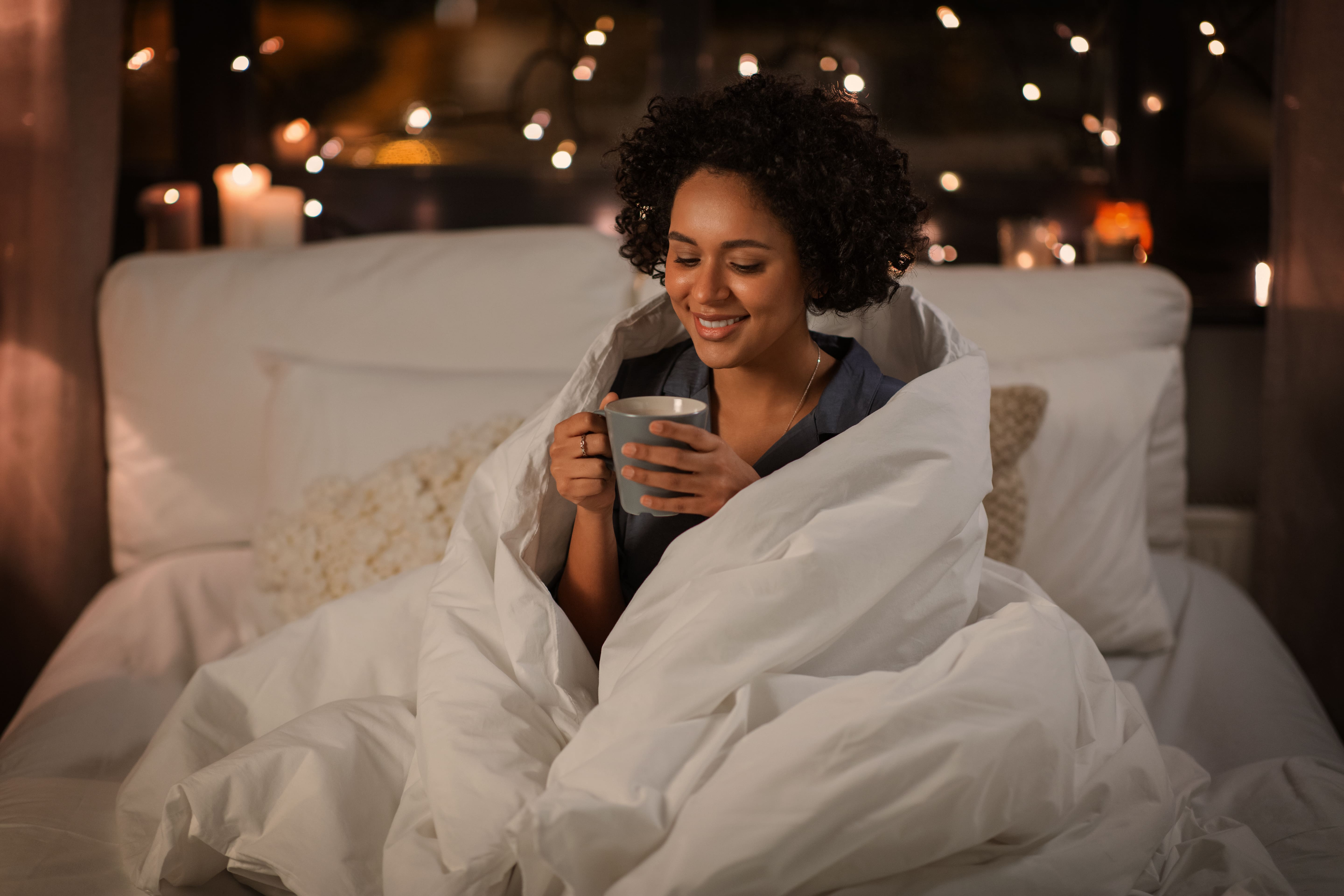 Woman sitting up in bed wrapped in a duvet holding a mug and smiling.
