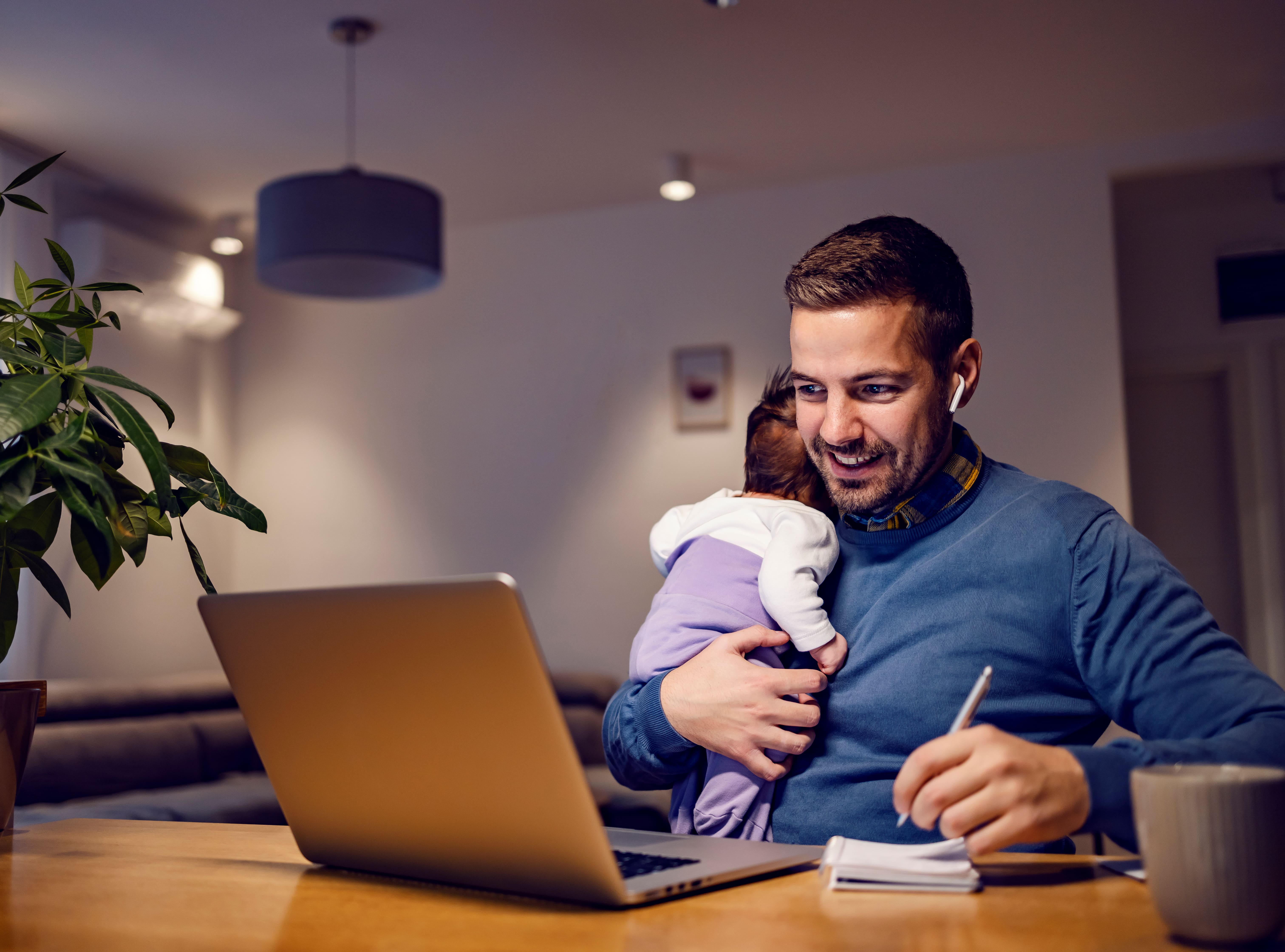 A dad working from home while comforting a new born baby as he takes meeting notes with a pen and paper.