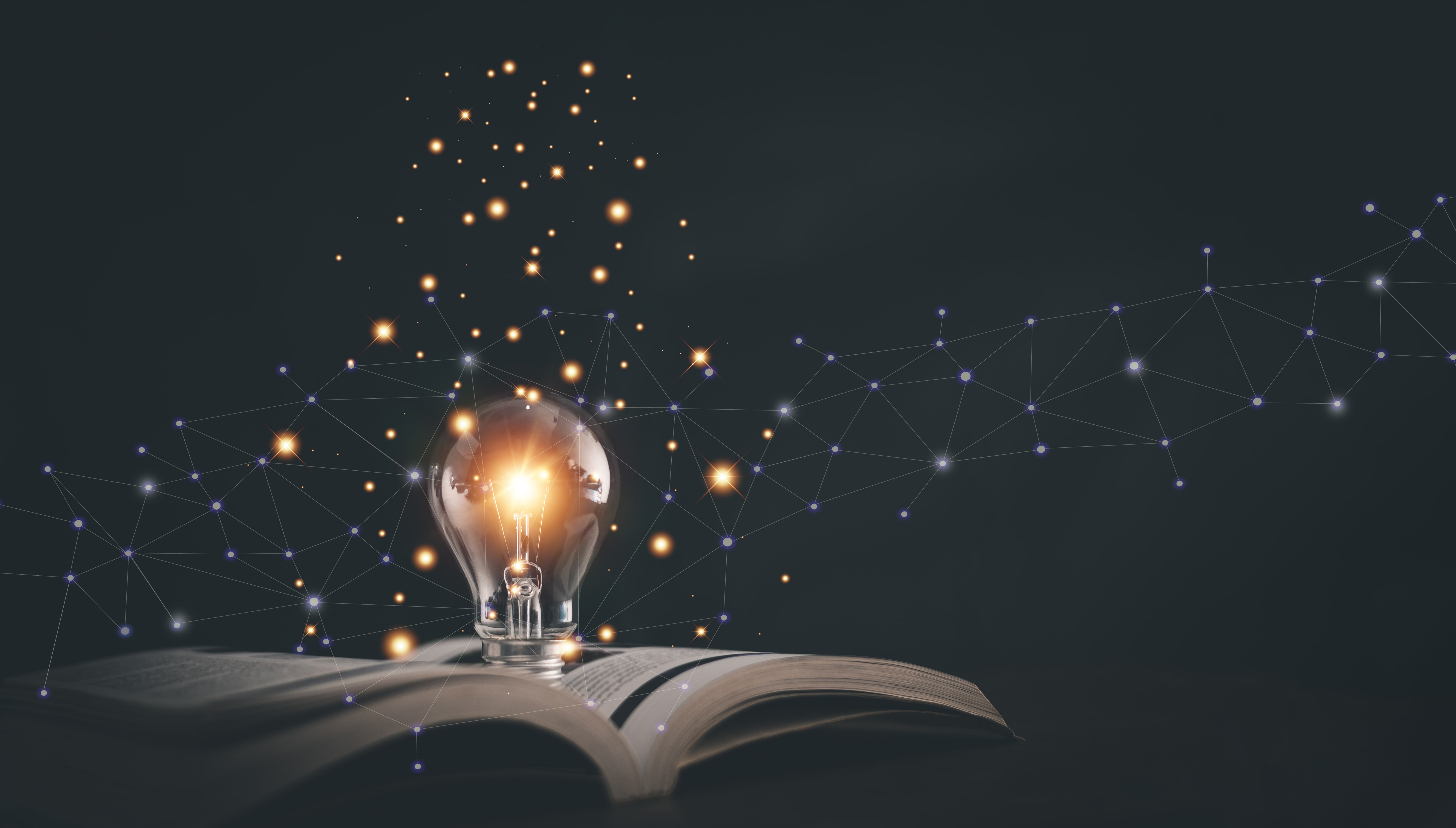 A glowing lightbulb sits on the open pages of a book and emits sprinkles of light. A constellation of light expands across the centre of the black background.