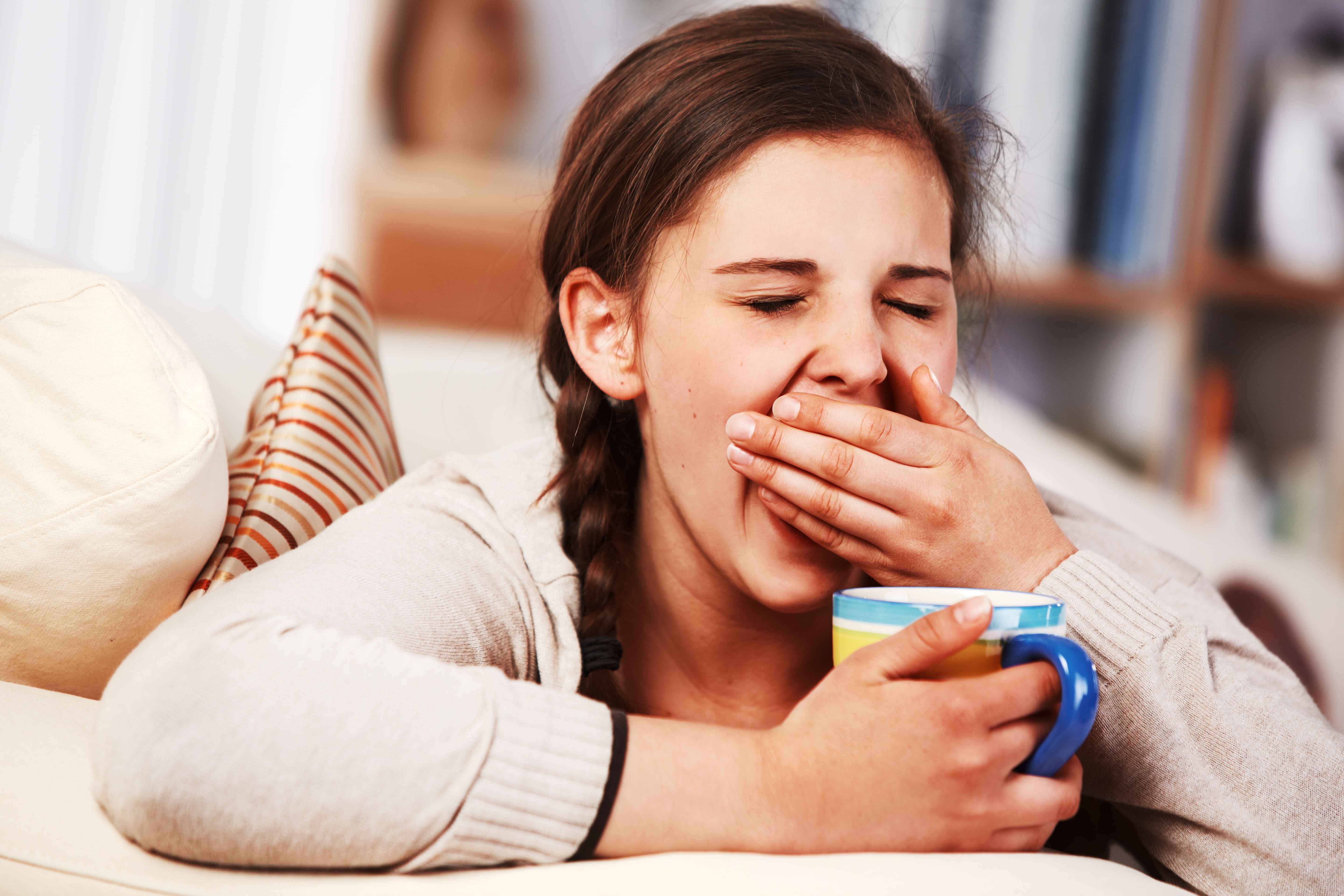 Teenage girl holding a cup in one hand and holding the other hand over her mouth as she yawns.