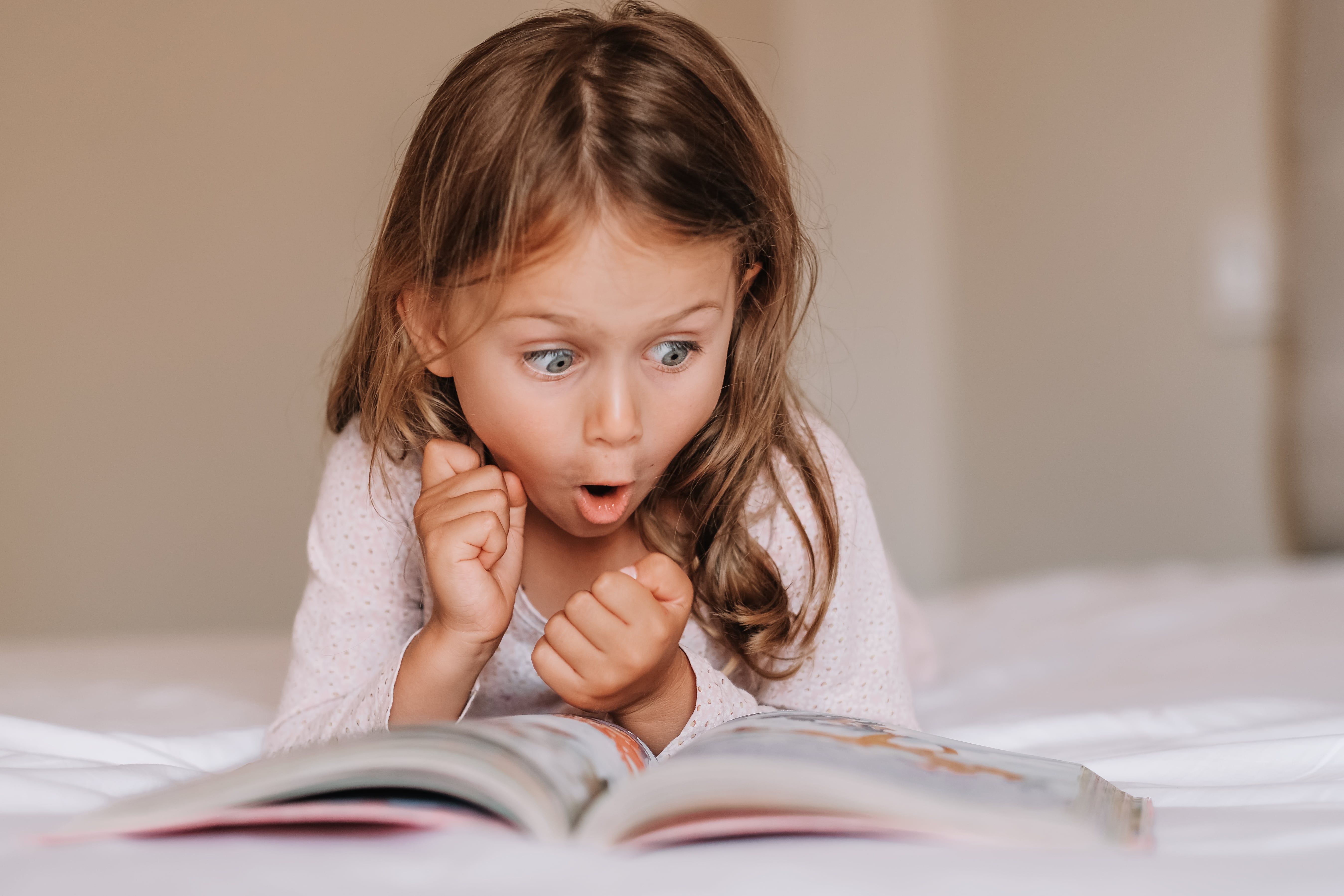 A little girl lying on her tummy in bed reading a picture book. She is completely surprised about what is happening in the story as her face is one of complete shock