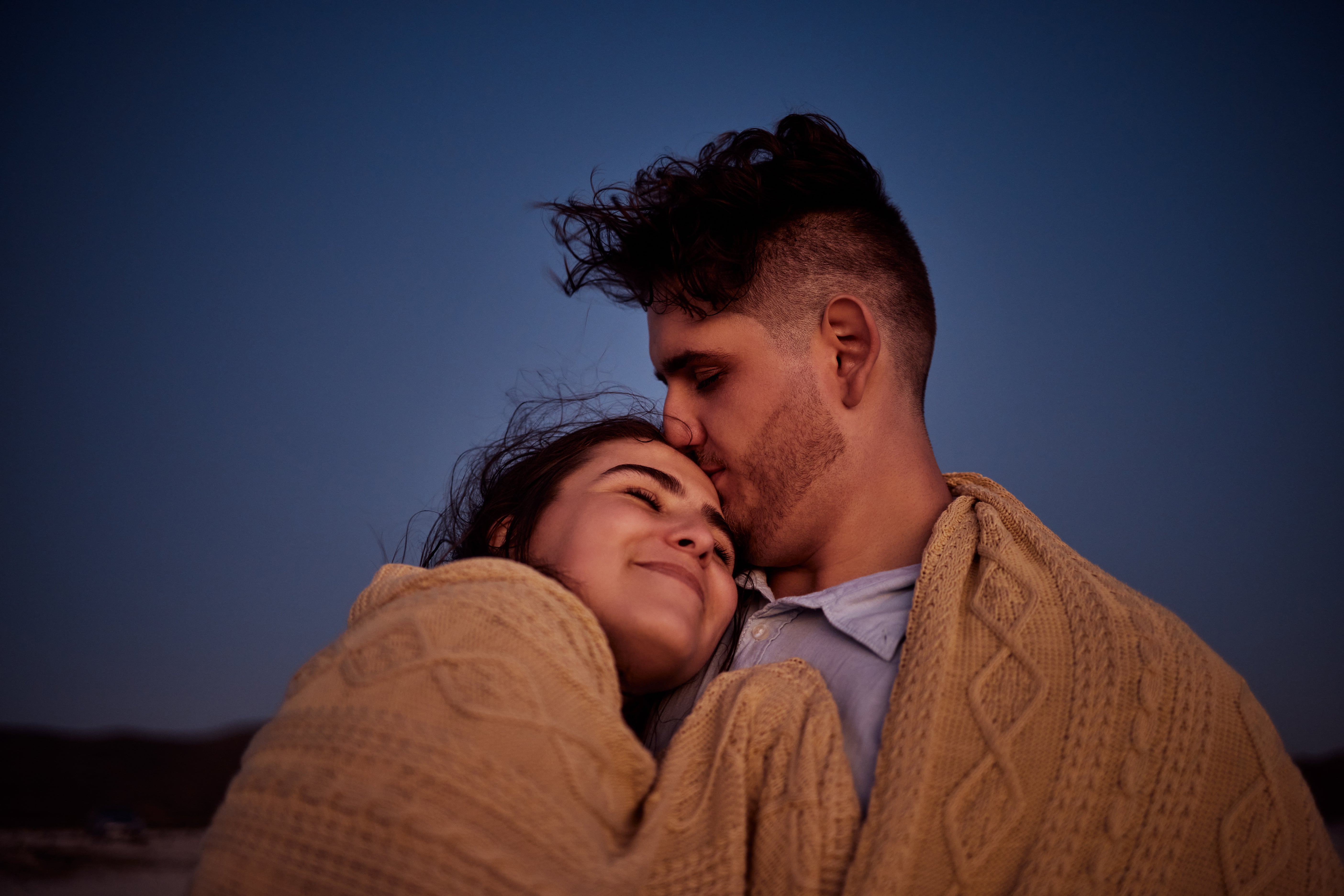 A couple embracing, wrapped in a blanket big enough for two as they star gaze outdoors.