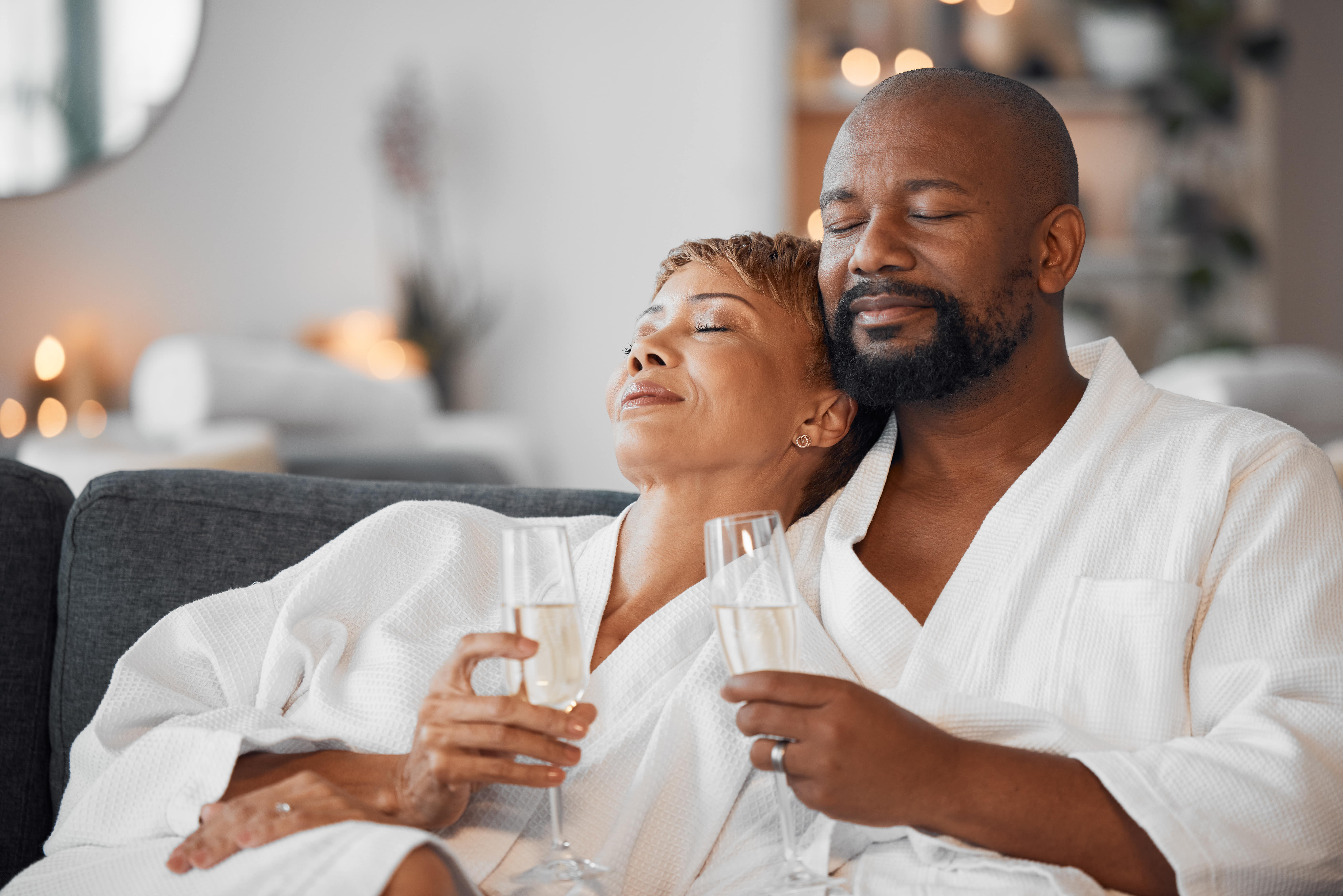 A couple wearing white spa robes, sipping champagne, and cuddling close together on the sofa in the comfort of their own home.