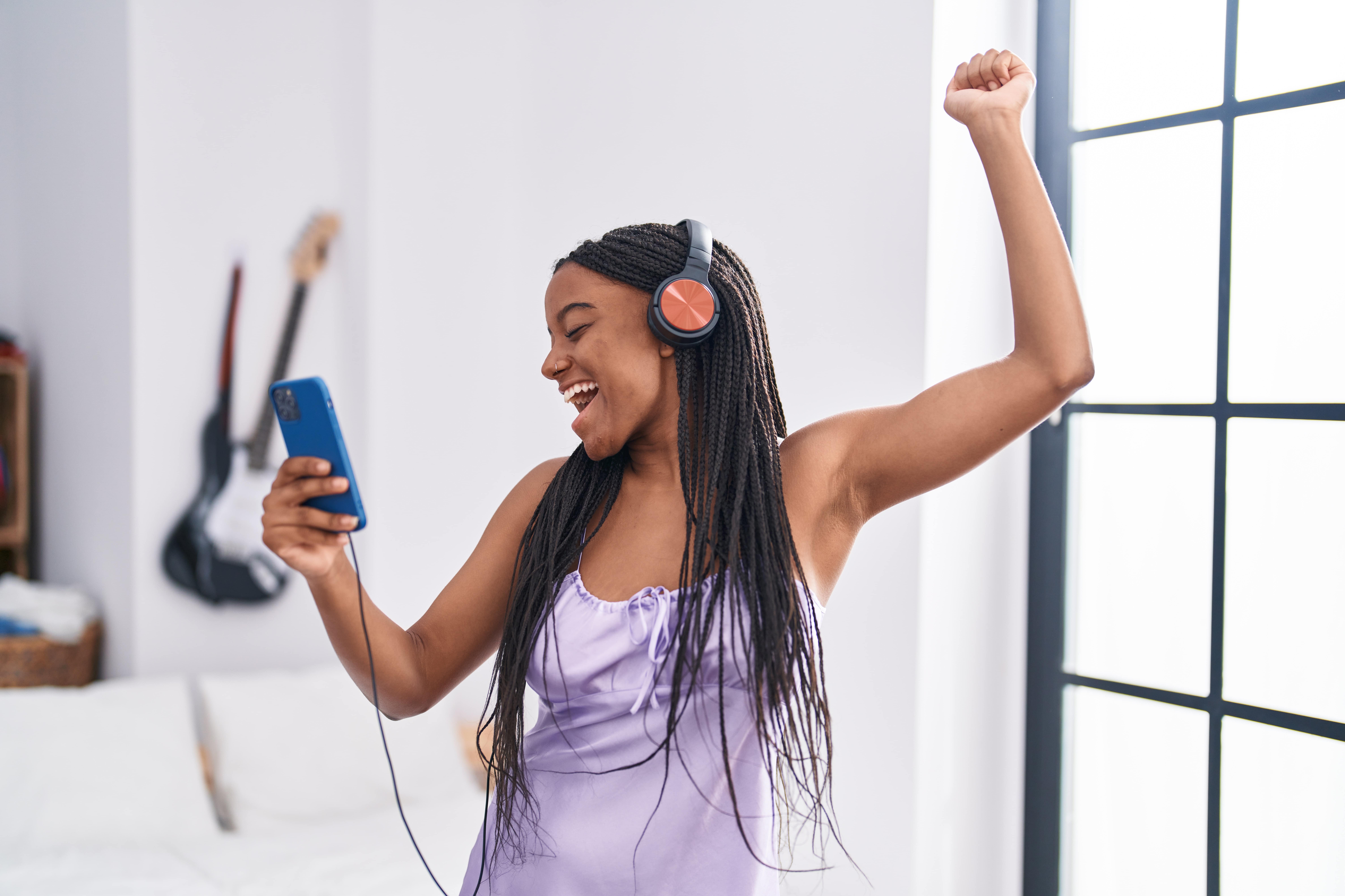 Woman dancing and smiling with headphones on listening to tunes from her mobile. An electric guitar hangs on the wall in the background.