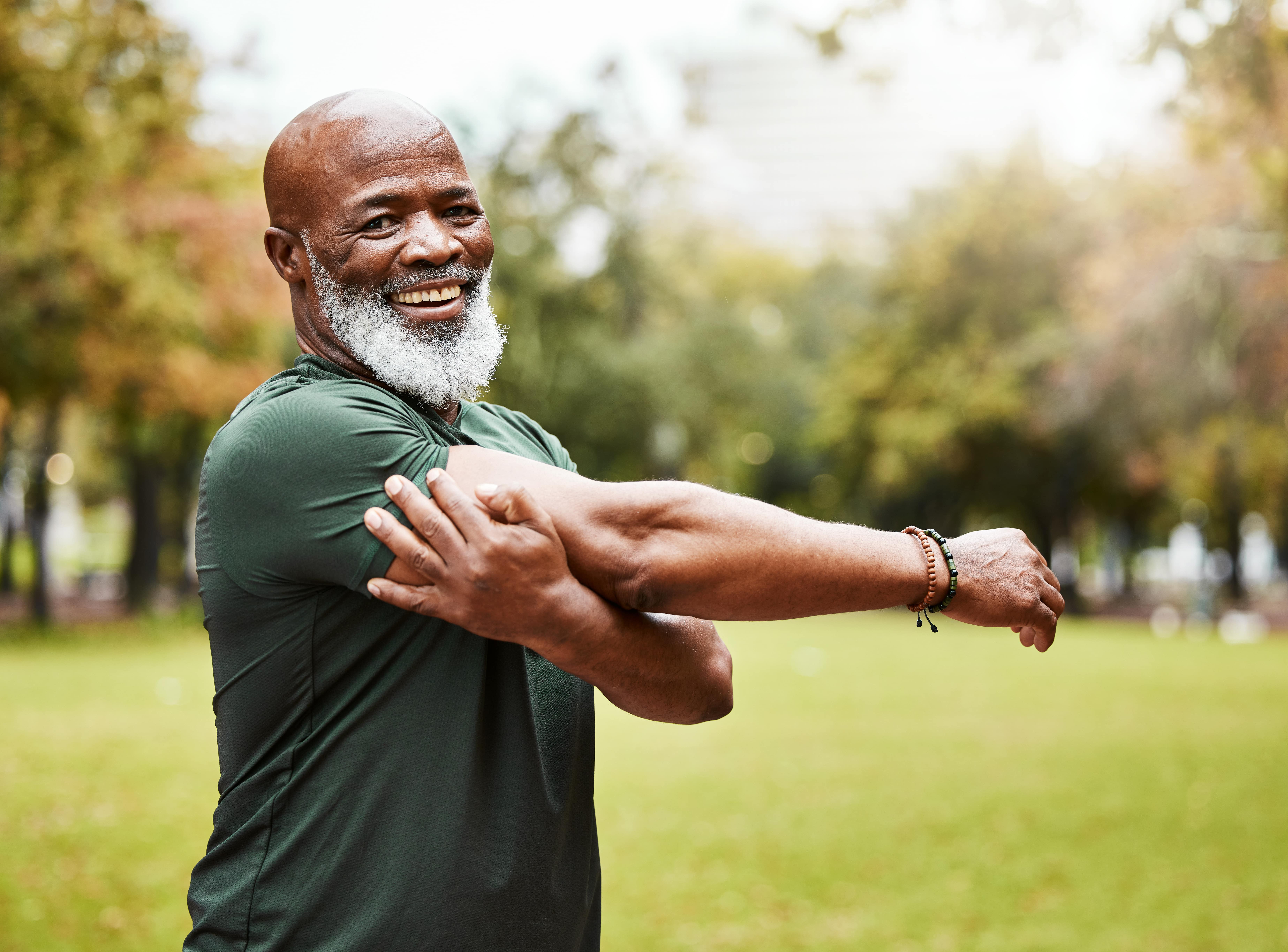 Man smiling in a park stretching his arms in preparation for exercise.