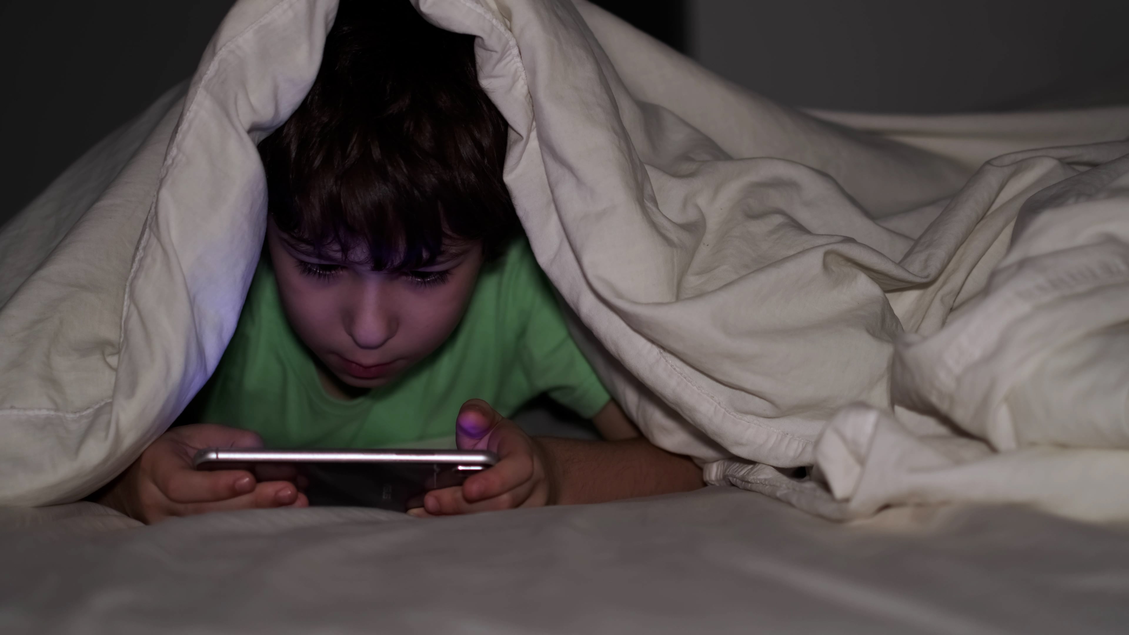Boy looks at a mobile phone under a duvet at night.