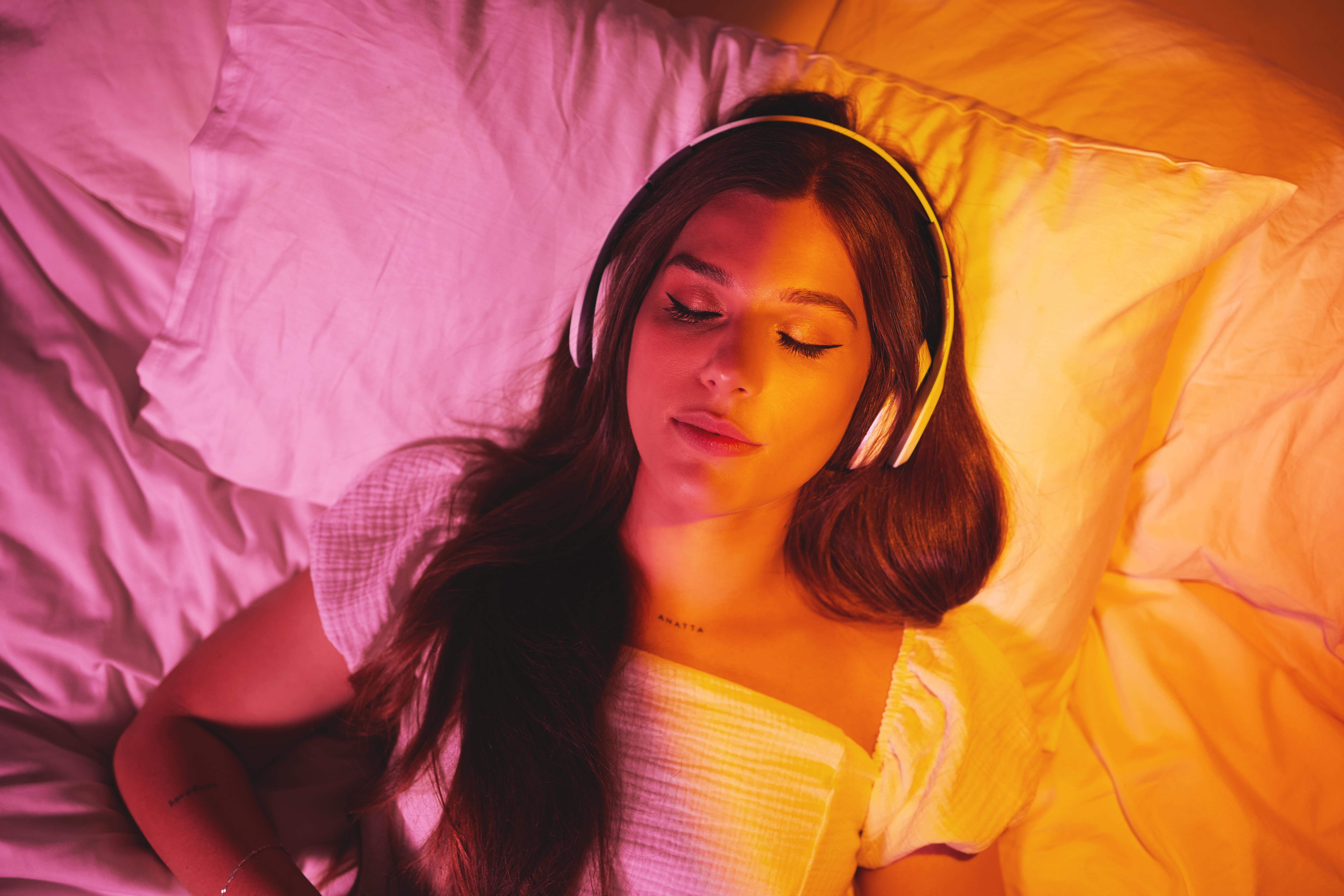 An aerial photo of lady lay on her back on her bed wearing headphones to listen to music while she sleeps. There is a pink and orange glow from lighting across the entire image.