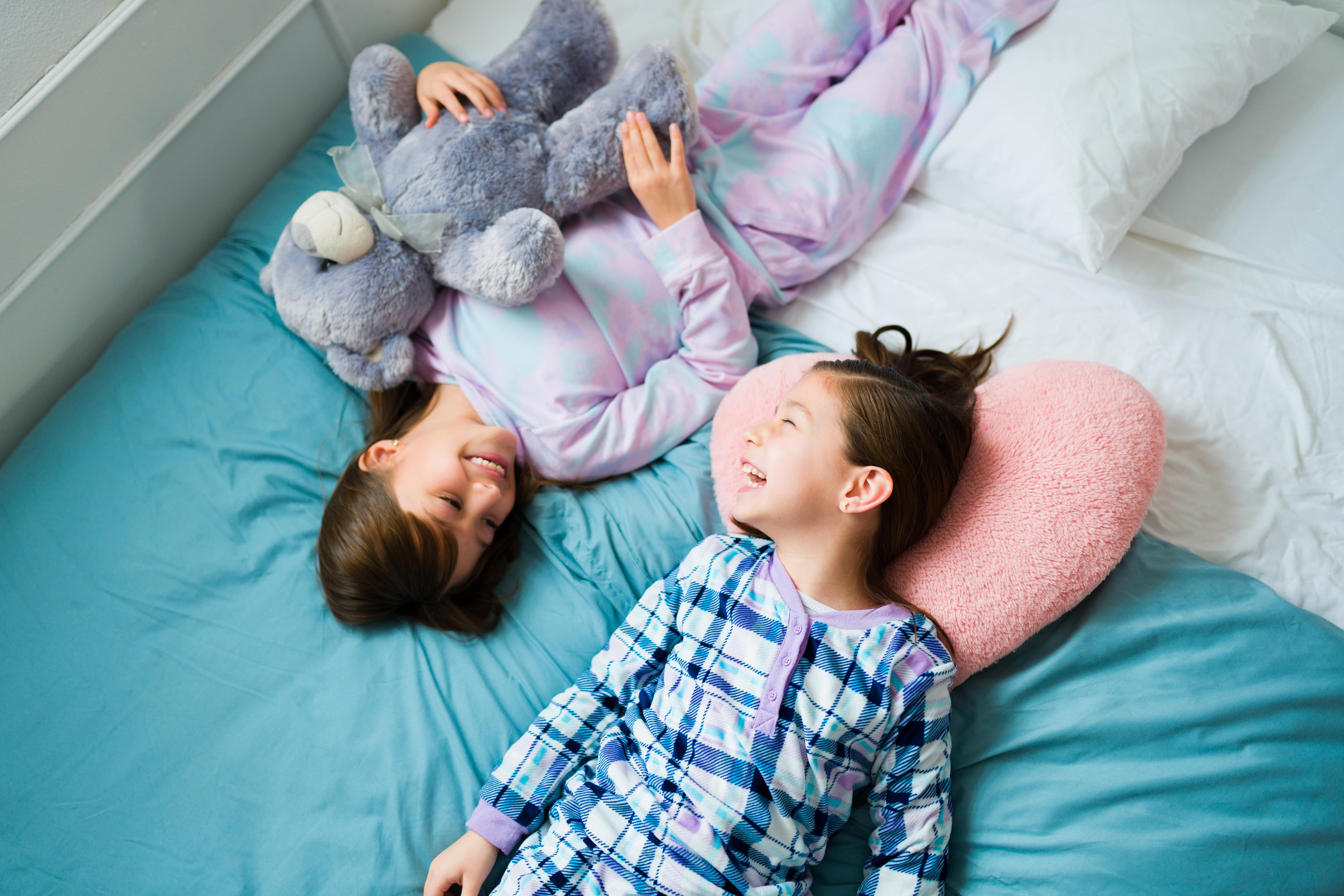 Two friends smiling in their PJs as they lie on a teal duvet. One is holding a fluffy purple teddy, the other is resting their head on a fluffy pink heart shaped pillow.