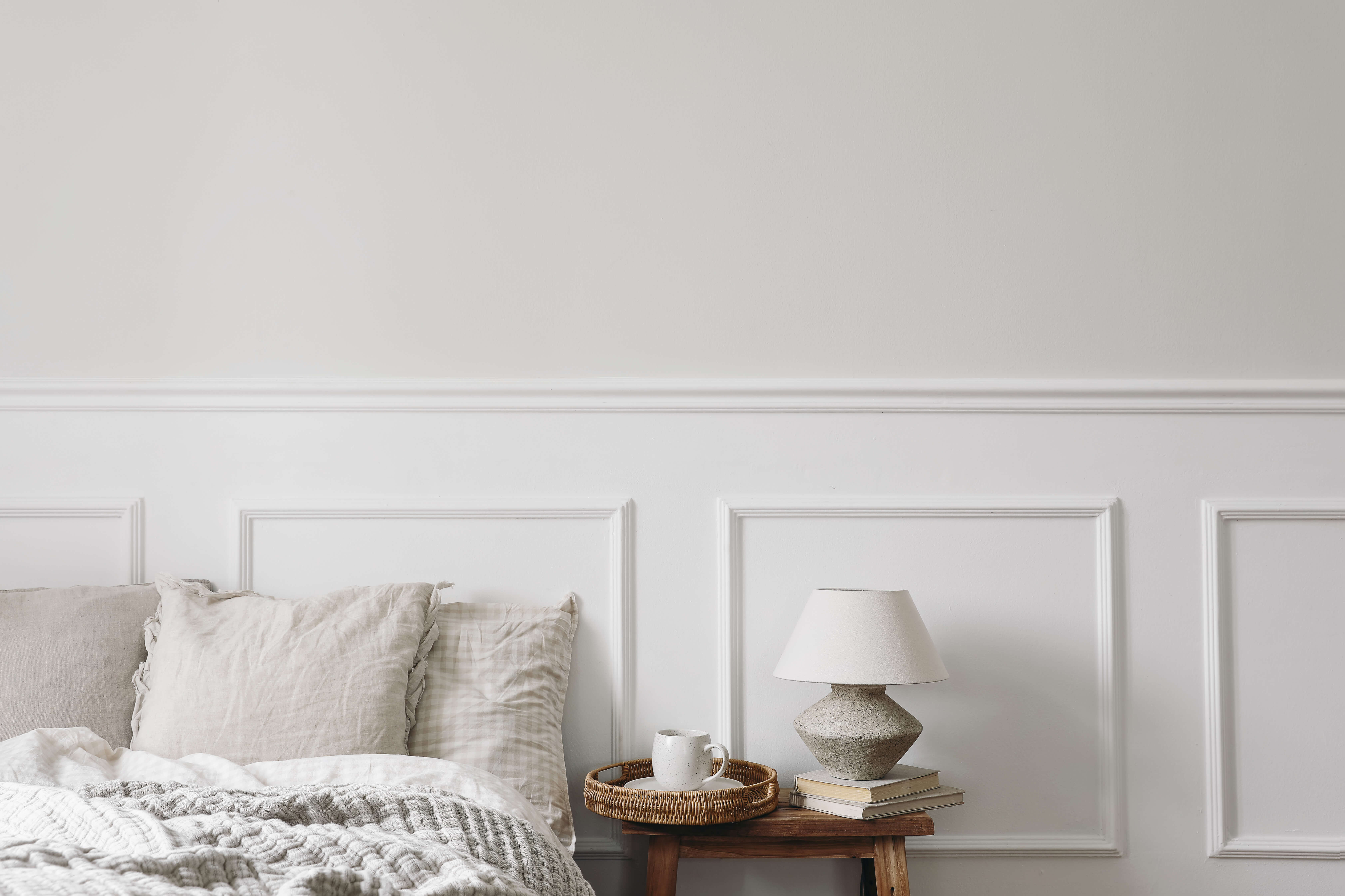 An empty bed with slightly ruffled covers set against a white half-panelled wall with a natural wooden bedside table featuring a pile of books, a lamp and a wicker drinks tray with a lone coffee cup and saucer