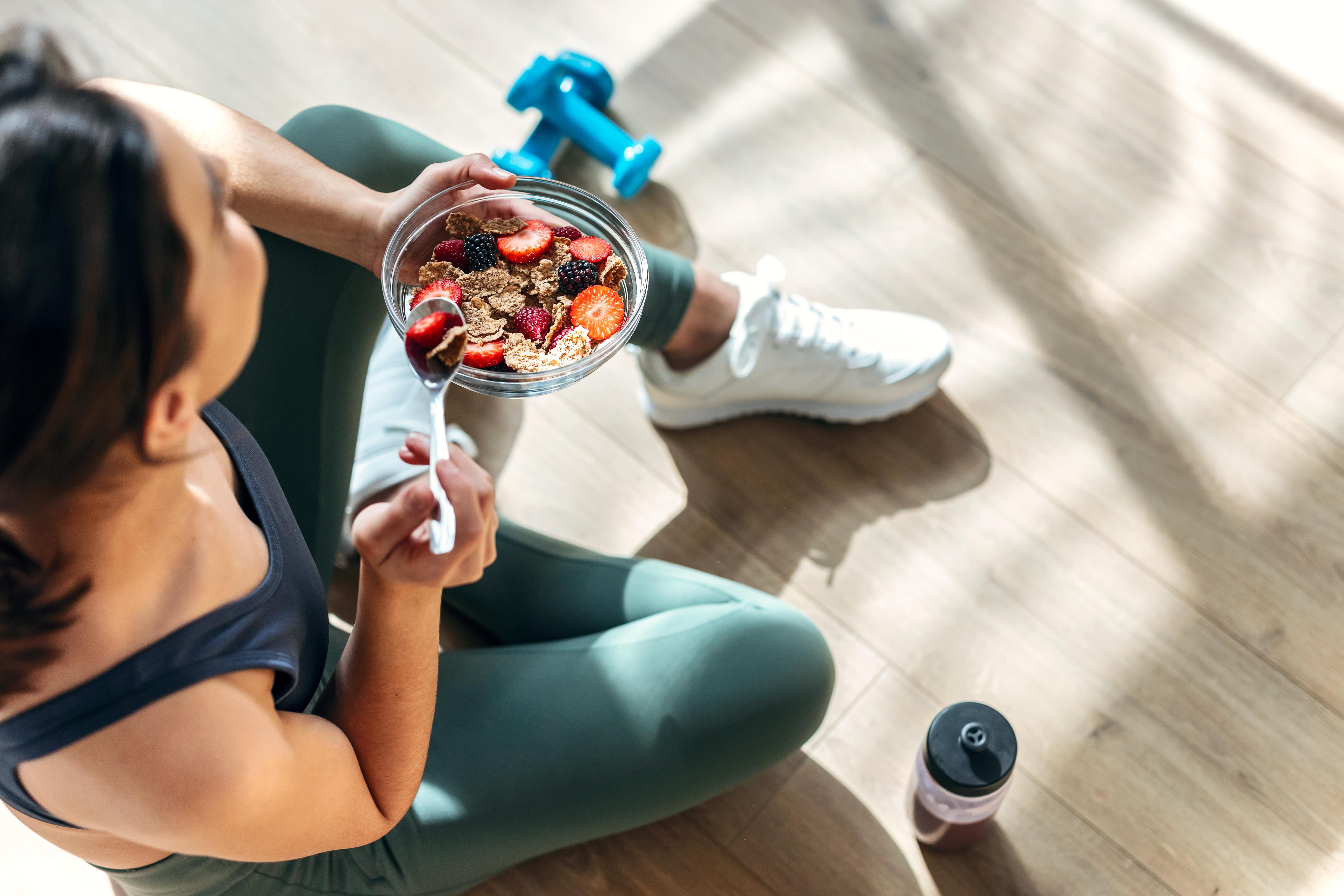 A woman eating a breakfast of berries and bran flakes after her morning exercise. Her weights and water bottle lie on the floor beside her
