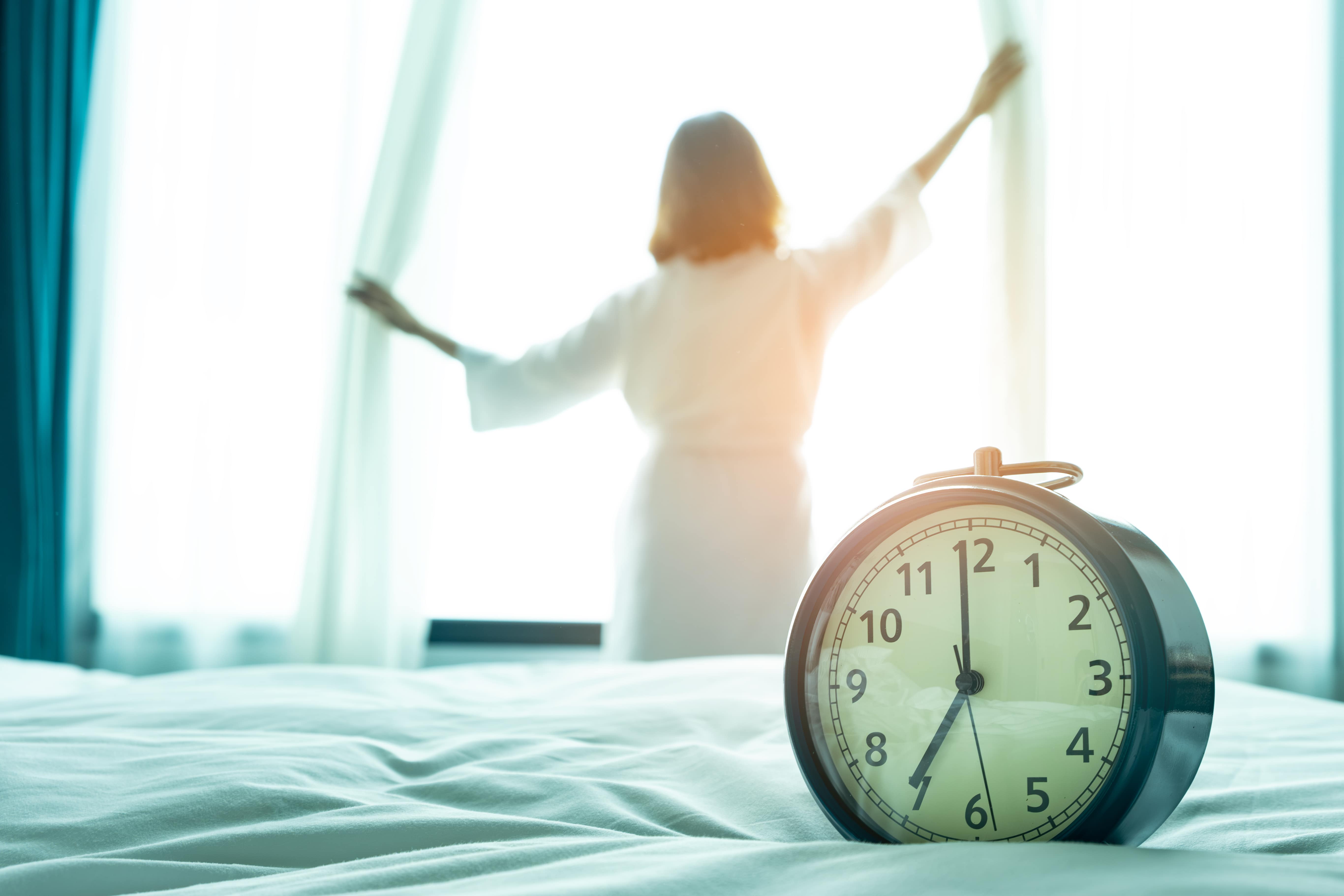 A woman opening the curtains at sun rise with a vintage clock in the foreground that reads 7am