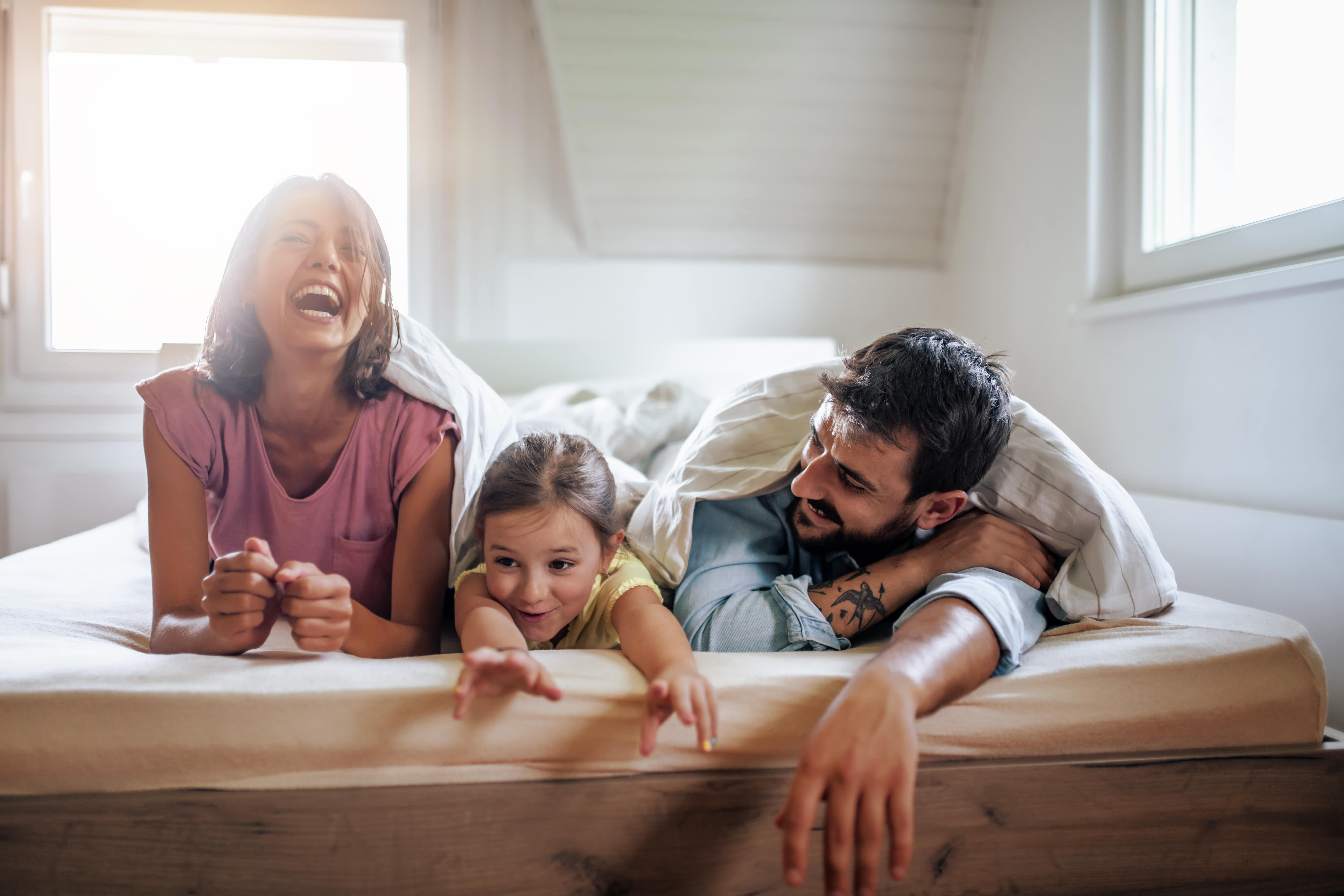 A family of three on a mattress smiling and having fun under the duvet.