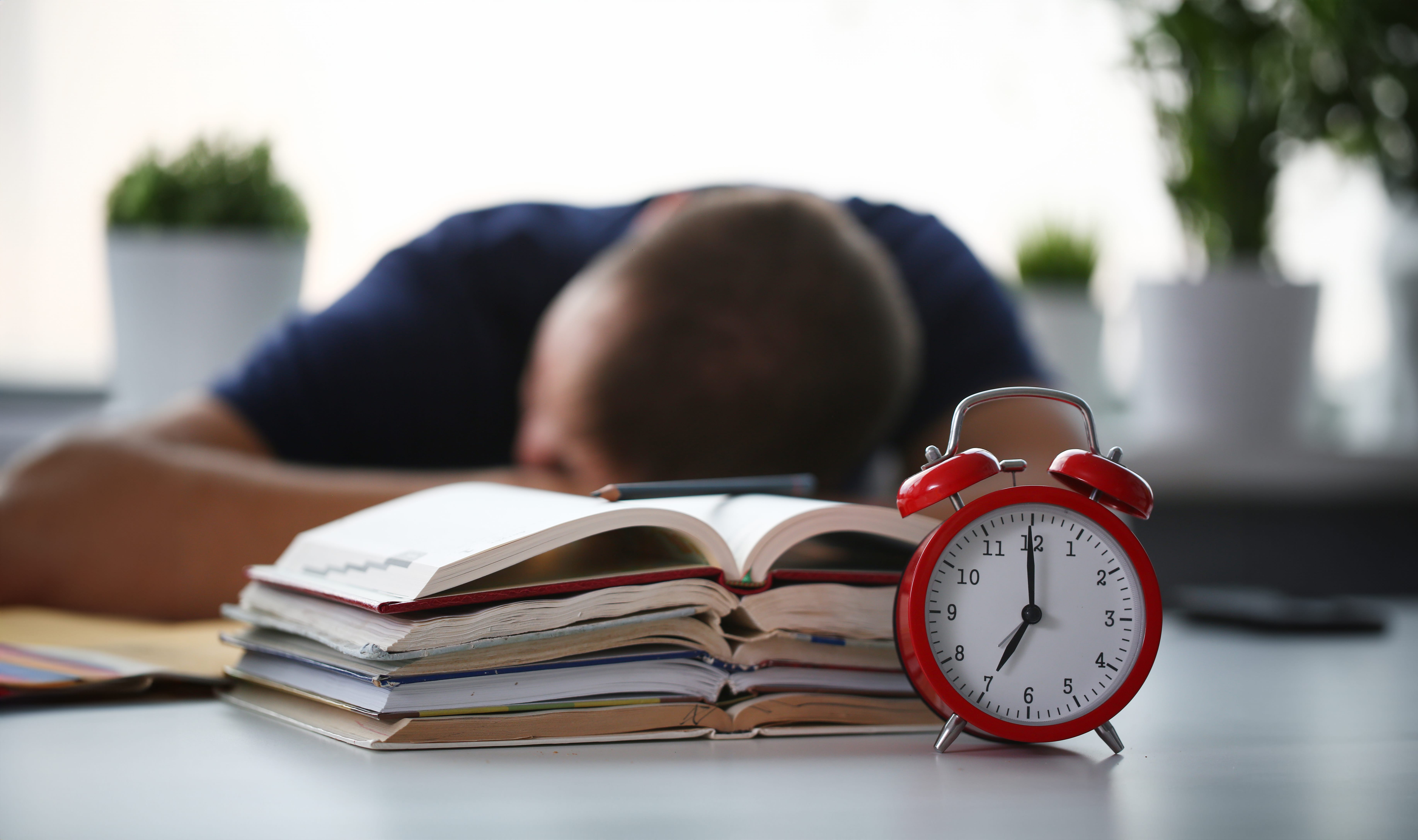 A student about to be woken up by his alarm clock after falling asleep at his desk with his head resting on a pile of books he was using for studying.