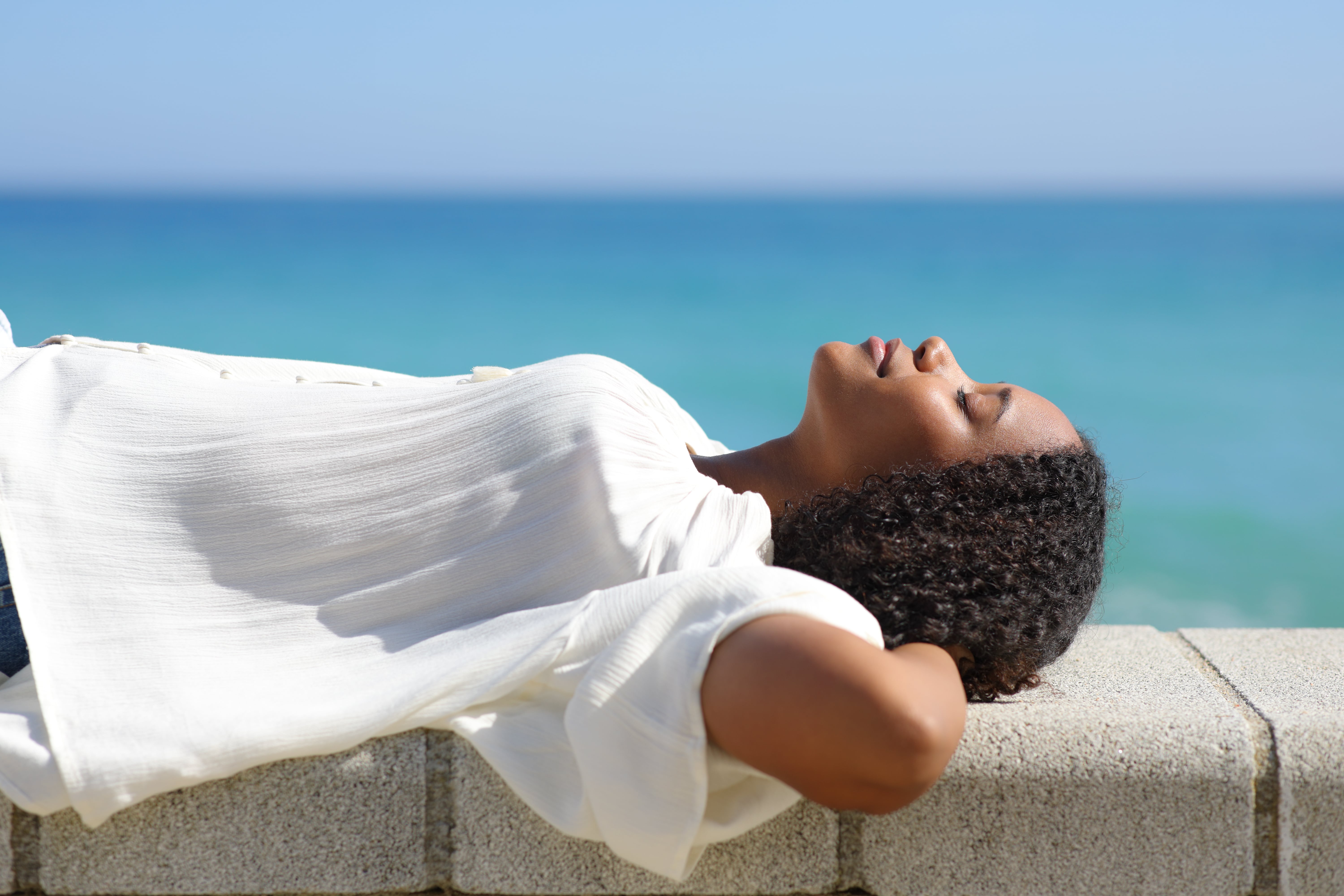 A woman lying on a wall with her eyes closed and her face to the warmth of the sun with the sea in the background