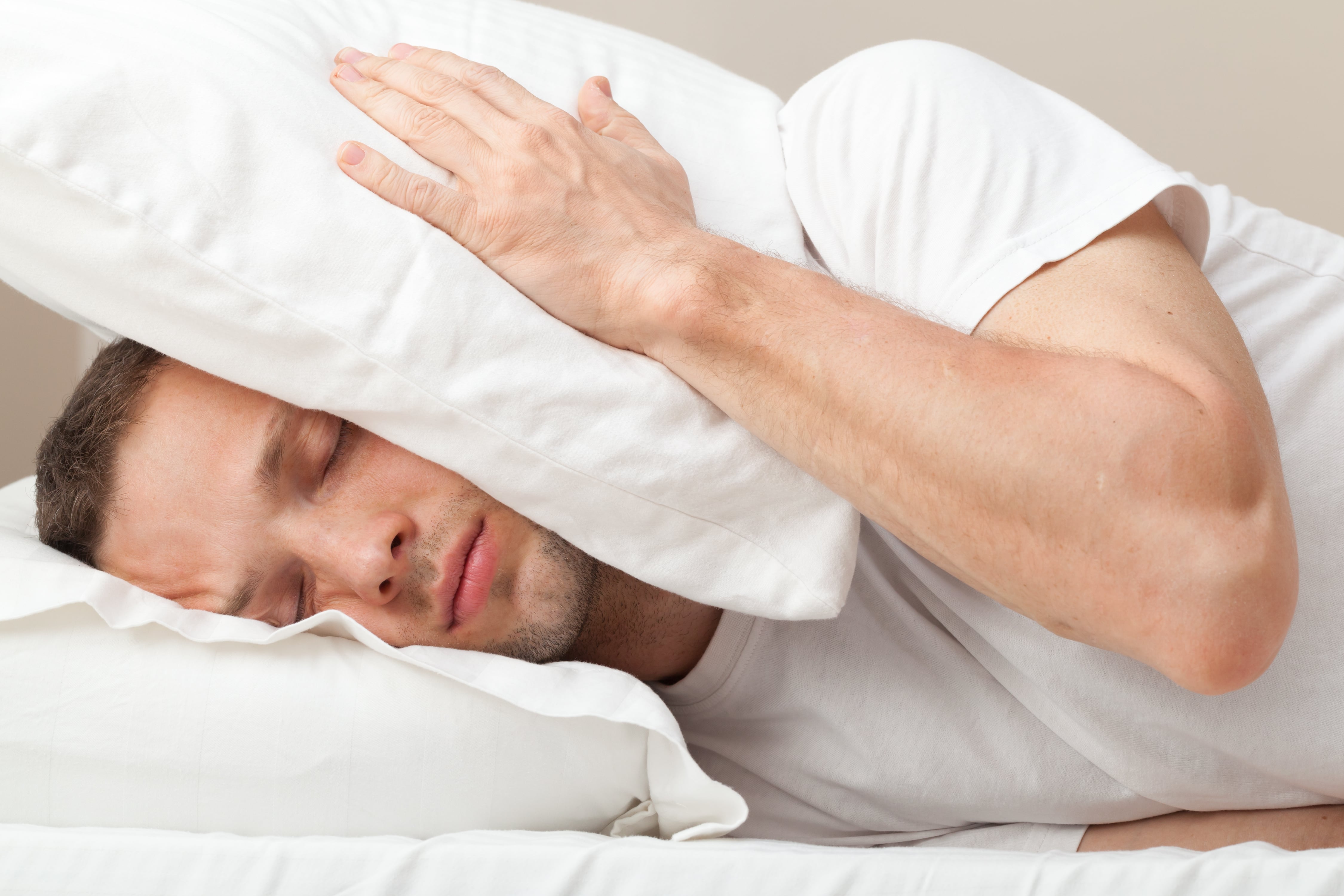 A man holding a pillow over his ears as he tries to block out noise so he can sleep