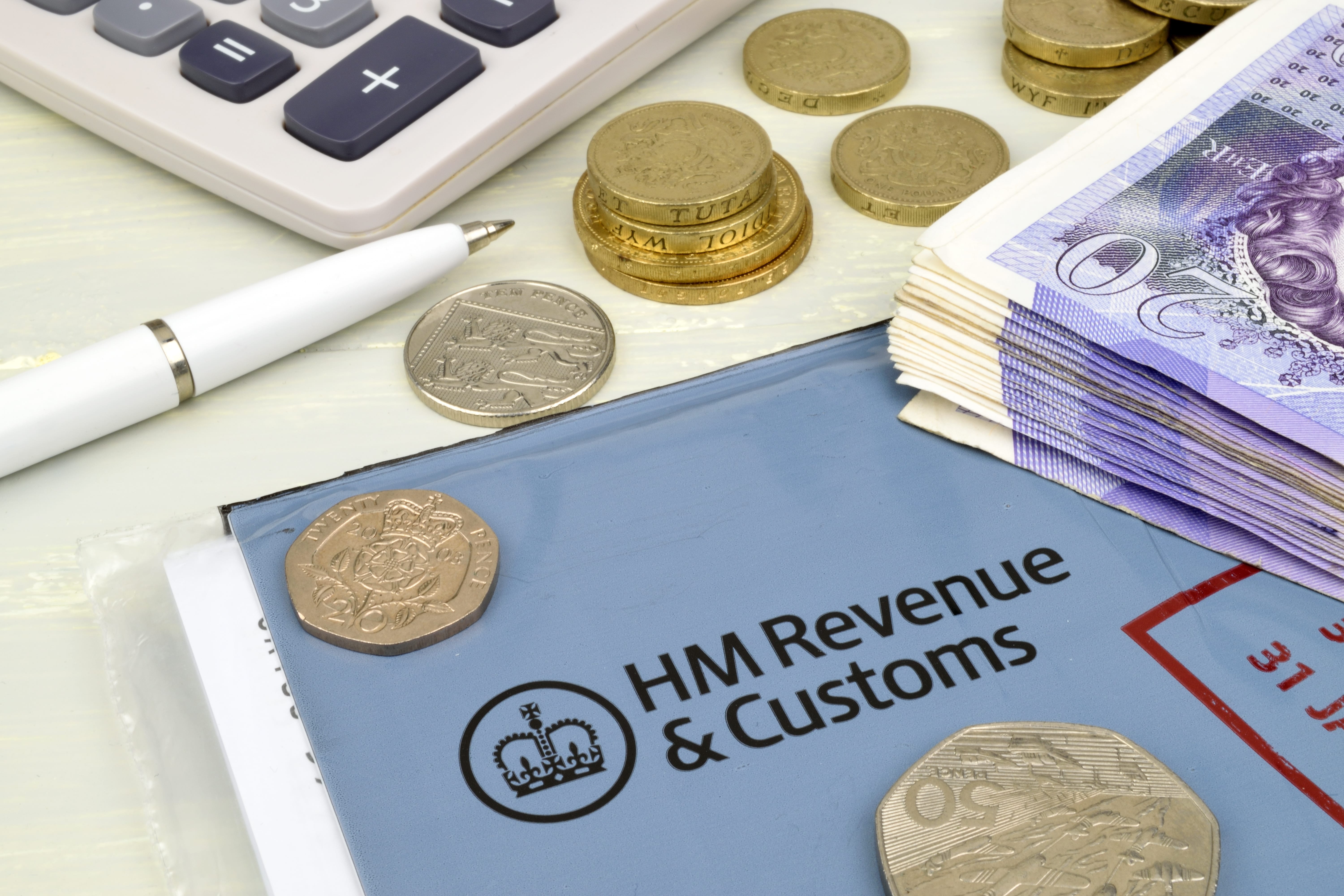 An HMRC document sits on the table in the foreground with a pile of £20 notes to one side and various denominations of coins strewn about. A calculator and pen sit on the table also.