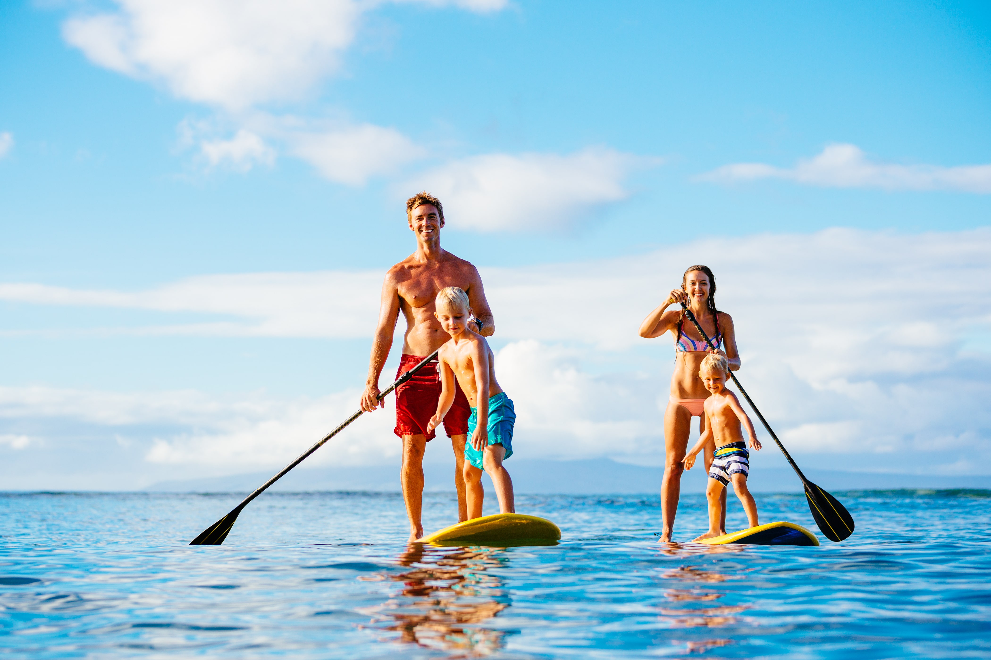 A family of four on paddle boards in the beautiful blue waters of the ocean. White fluffy clouds hang in the sky.