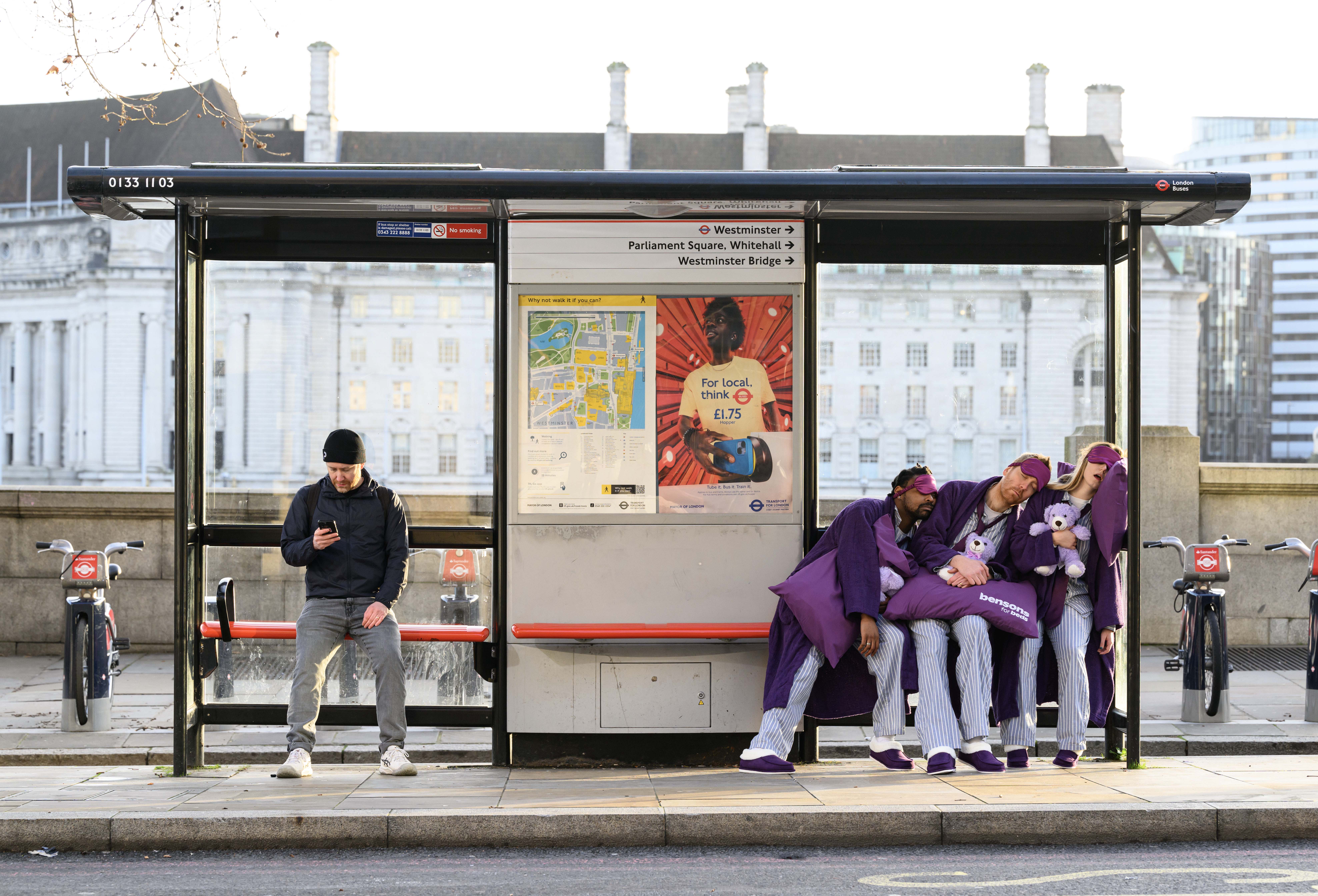 Three people asleep at a bus stop wearing pyjamas, purple dressing gowns, and purple slippers. Each has their own Bensons for Beds pillow, sleep mask, and teddy bear. A man sits solo at the other side of the bus stop busy on his smart phone as he waits for a bus to work.