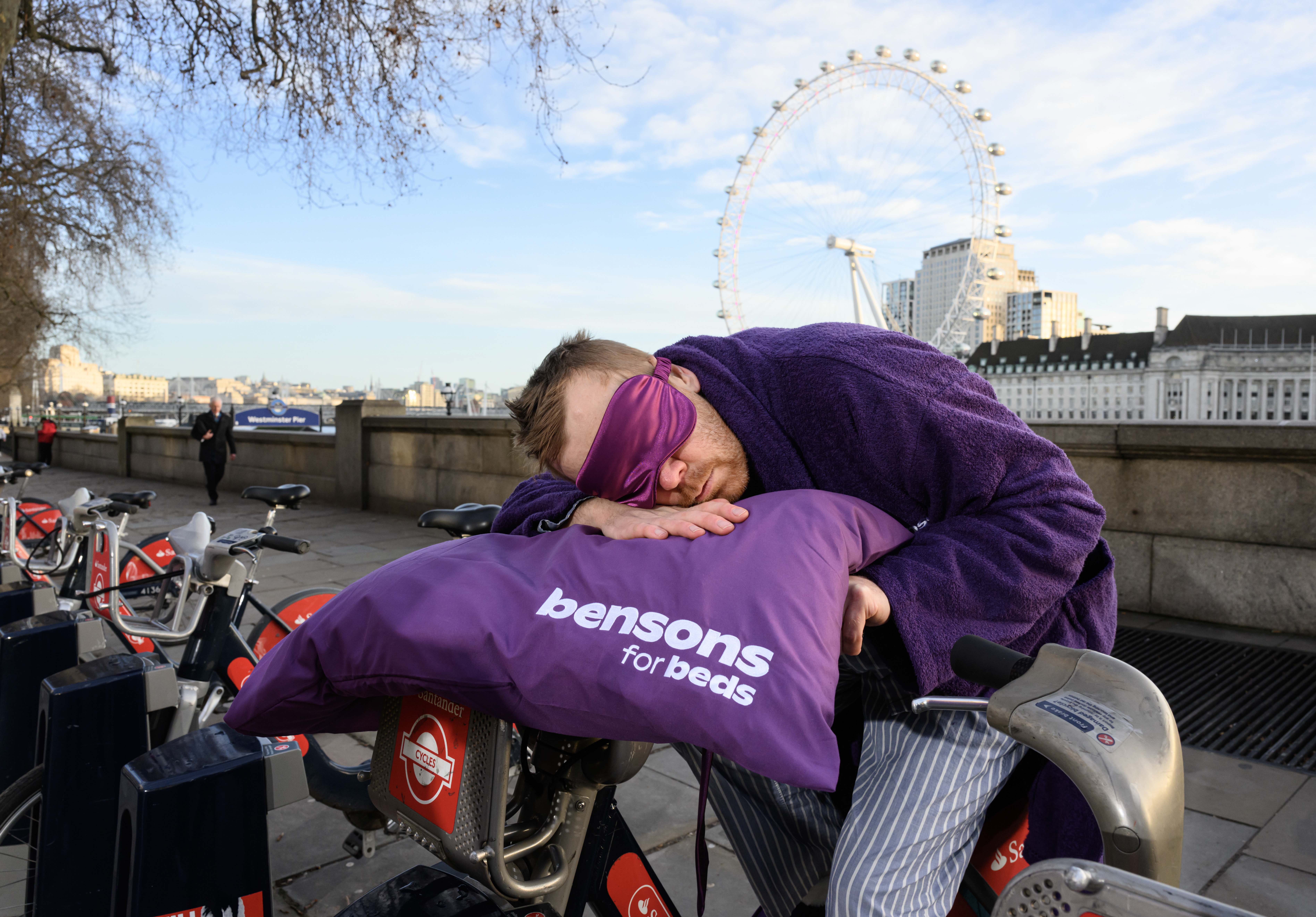 A man asleep on a parked bike for hire. He is wearing a purple dressing gown, pyjamas, and a sleep mask and in the background the London Eye is visible.