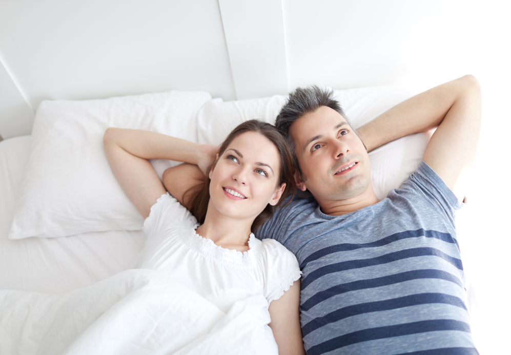 A smiling man and woman lie close to one another in bed and look to the ceiling.