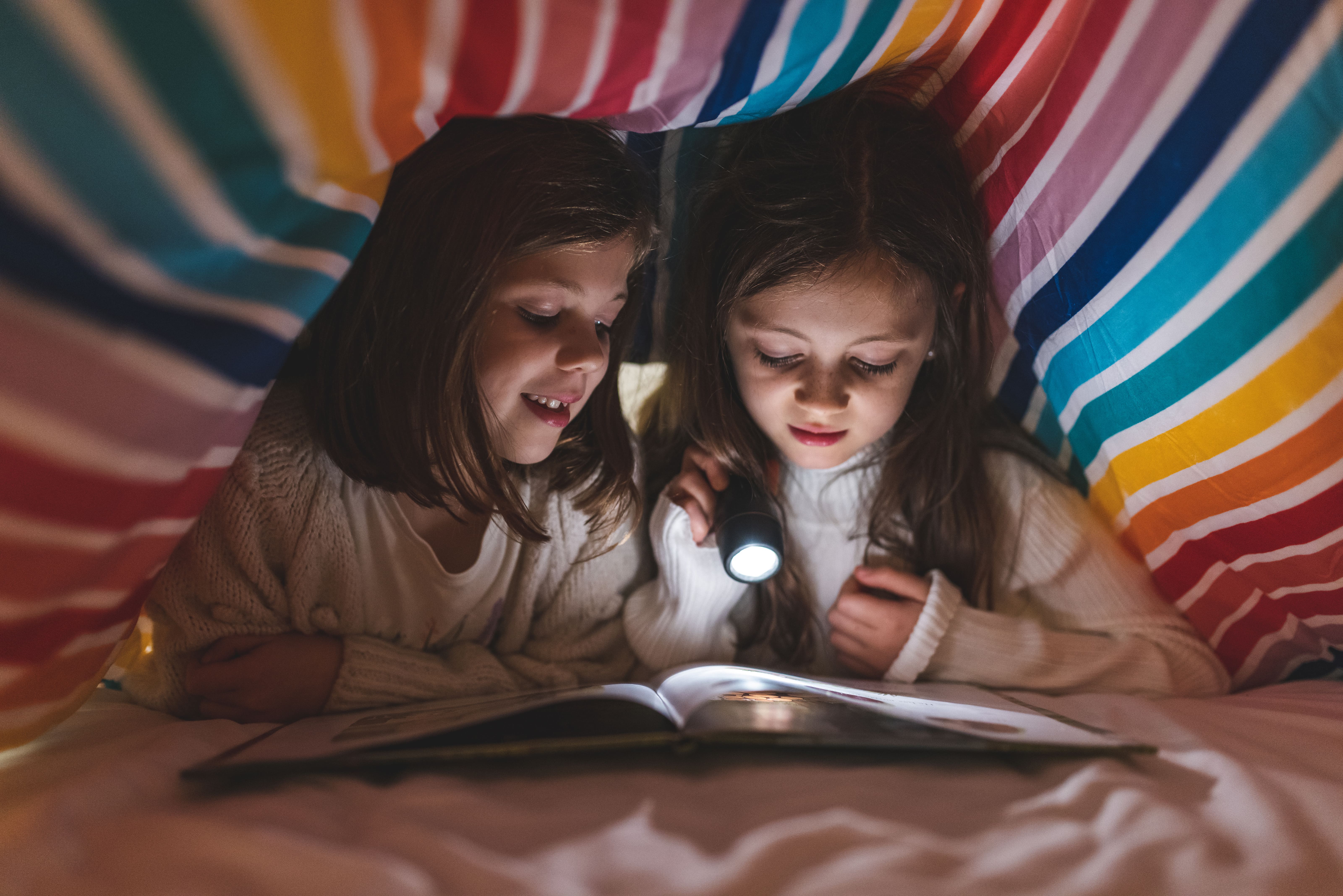 Two young sisters hiding under a striped duvet reading a book with a torch