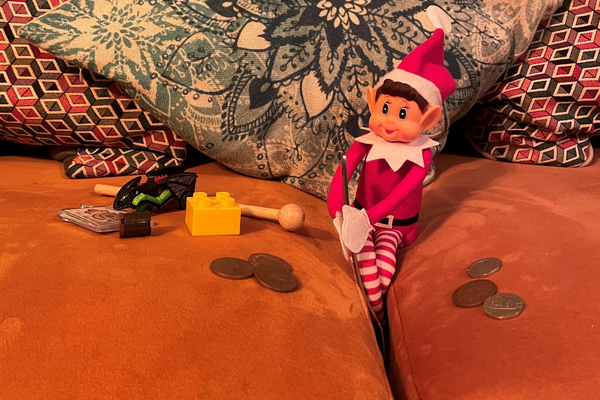 The elf on the shelf digging in between the couch cushions for treasure. A pile of coins and toys lie on either side of the elf