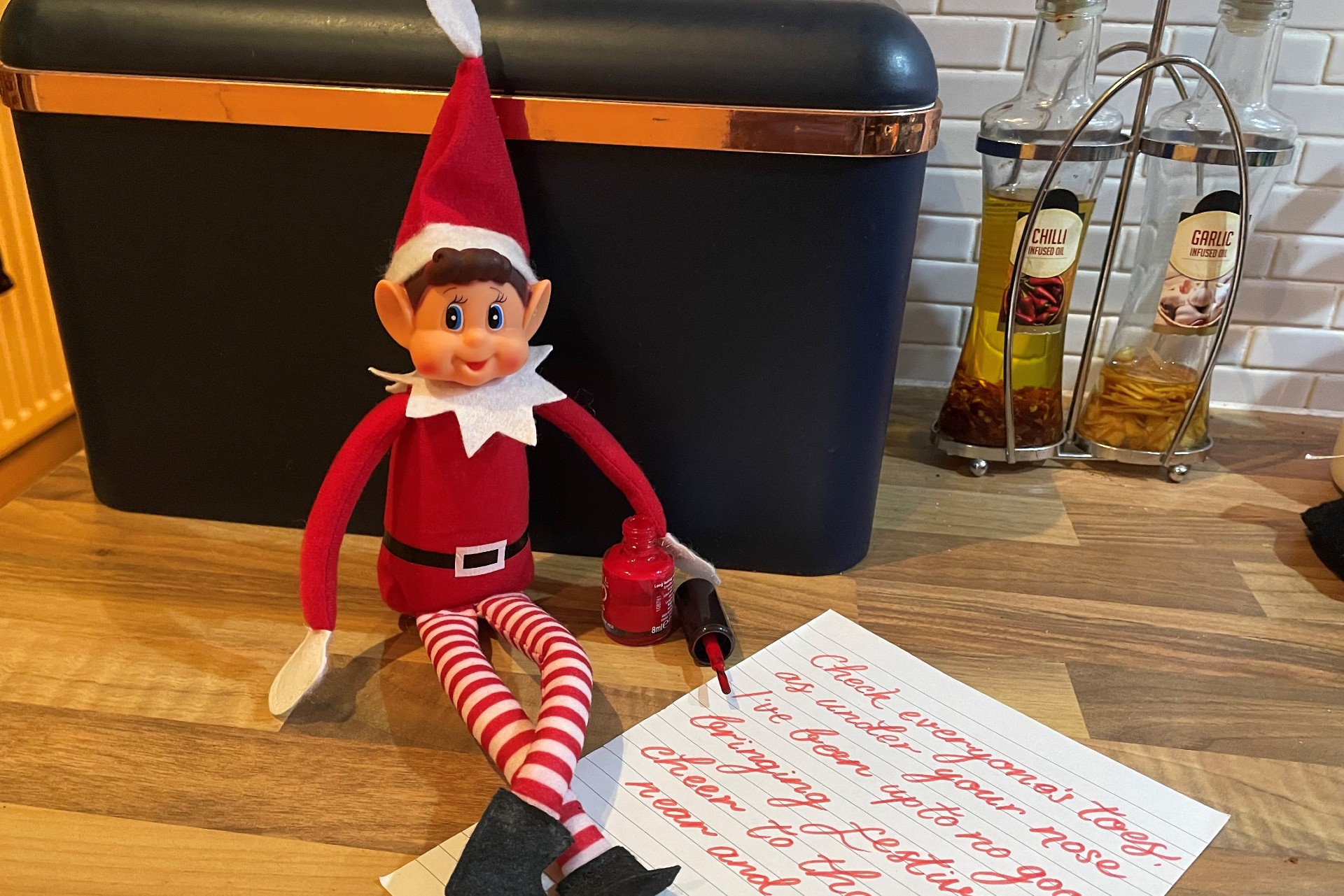 An elf on the shelf sat with a note and an open bottle of red nail varnish