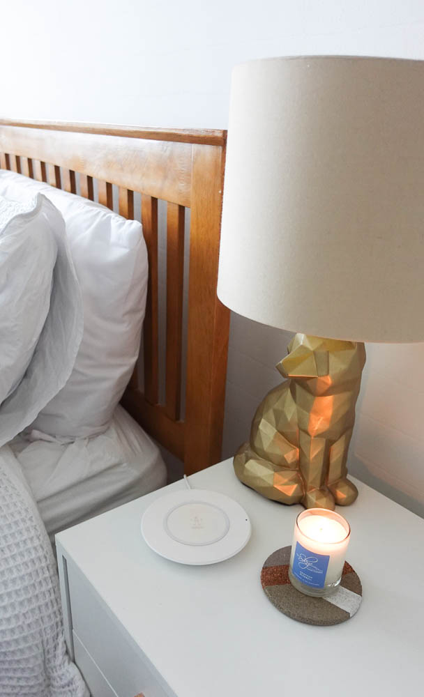 Two pillows are propped up against a wooden bed frame with a slatted headboard. A lit candle and a lamp on a set of bedside drawers are in the foreground.