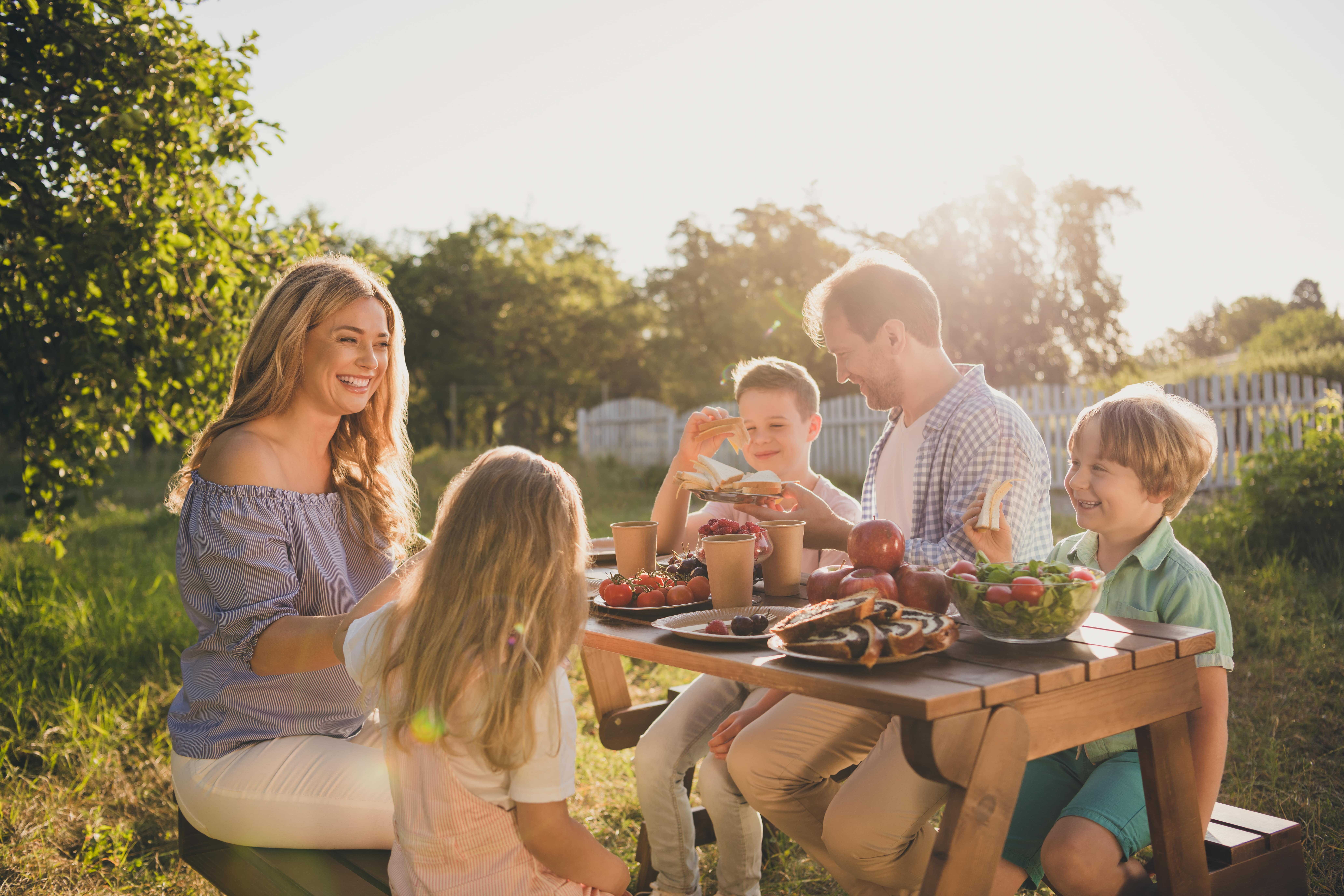 A family of five sat in the the garden surrounded by greenery eating a lunch of sandwiches, salad and cake at a picnic table