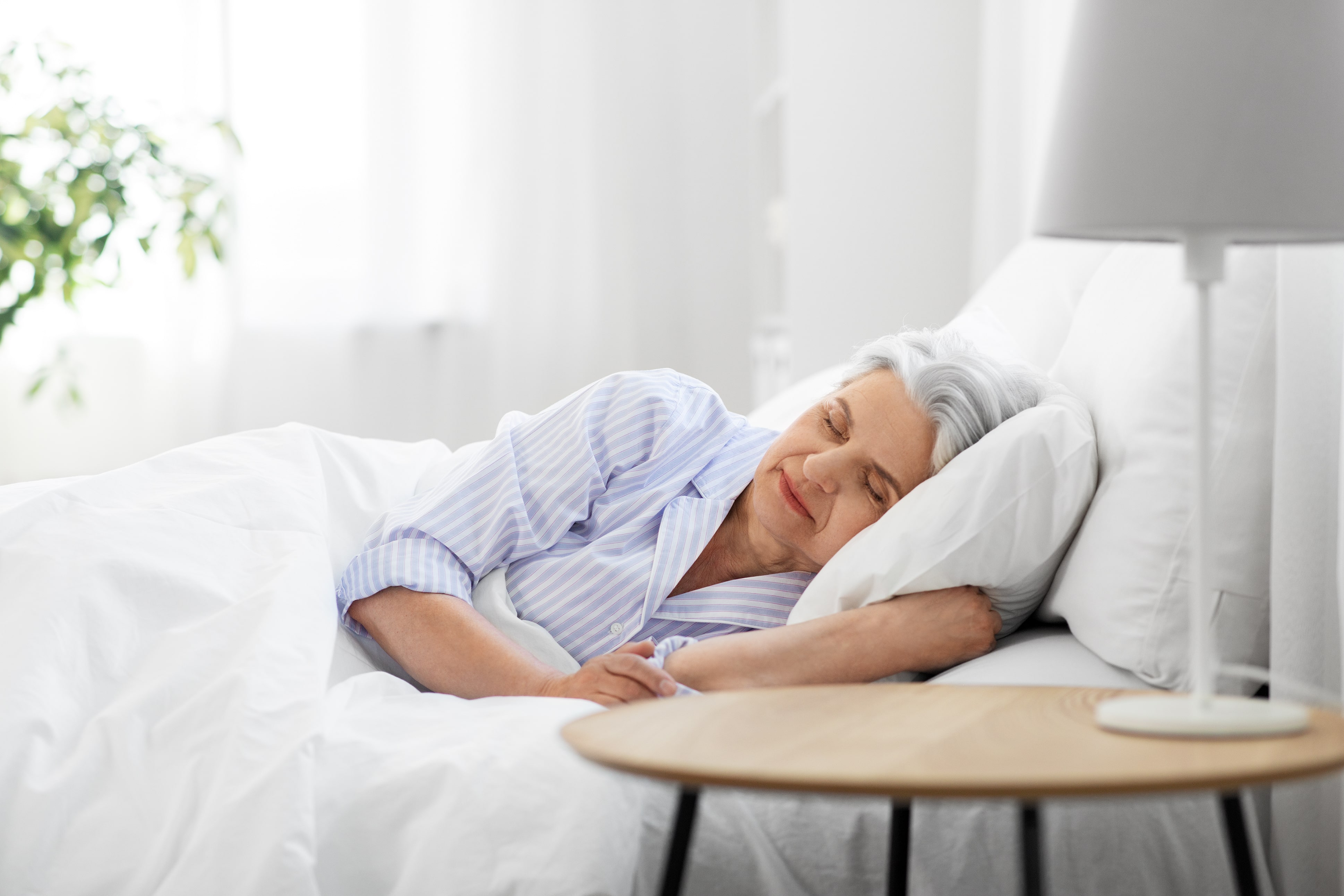 An older lady asleep on her side in bed and in the background is a green-leafed houseplant