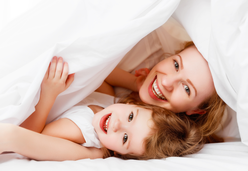 Woman and young girl smile to the camera as they lie beath a sheet.